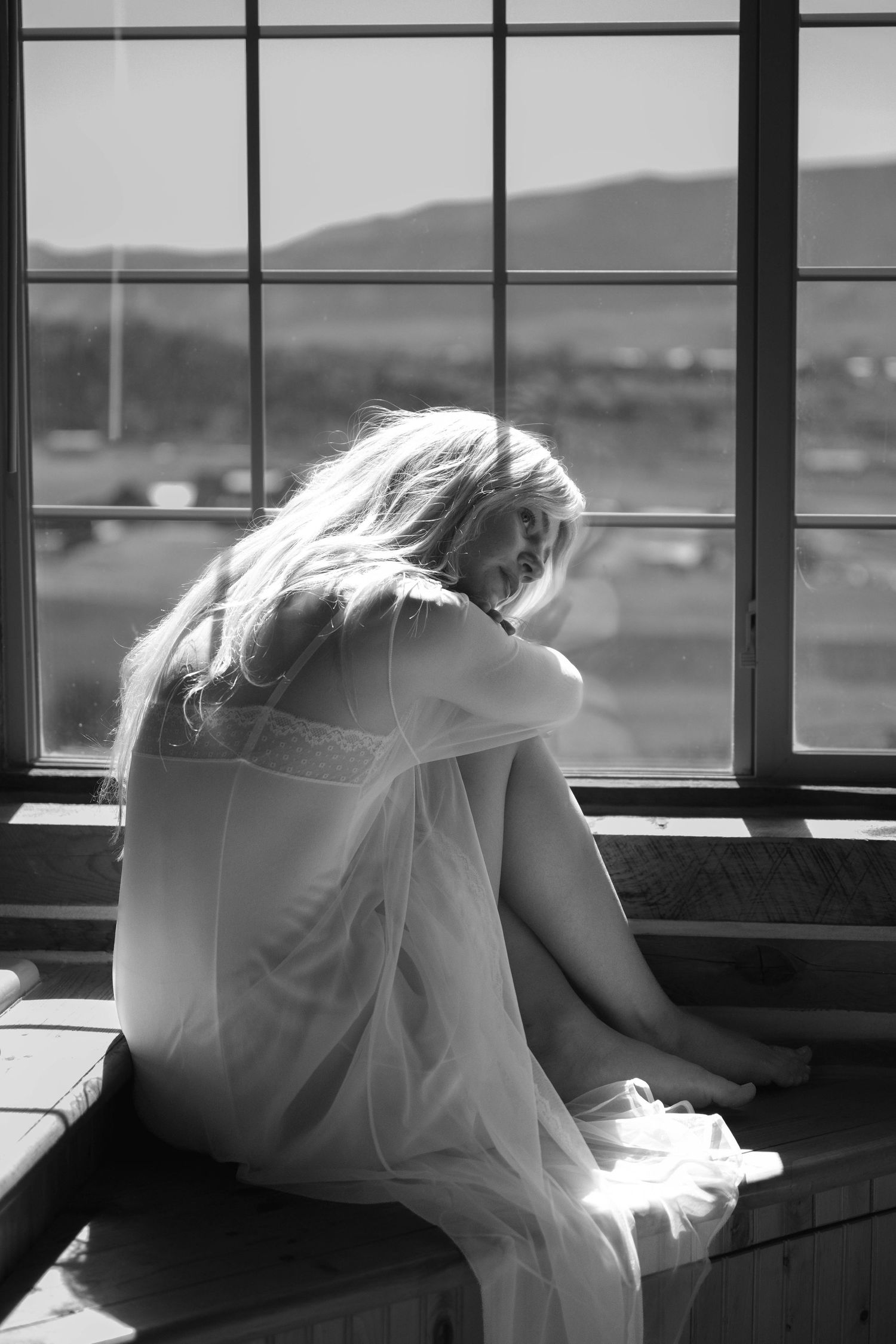 Blonde woman in white dress sitting alone on window sill in black and white photo.