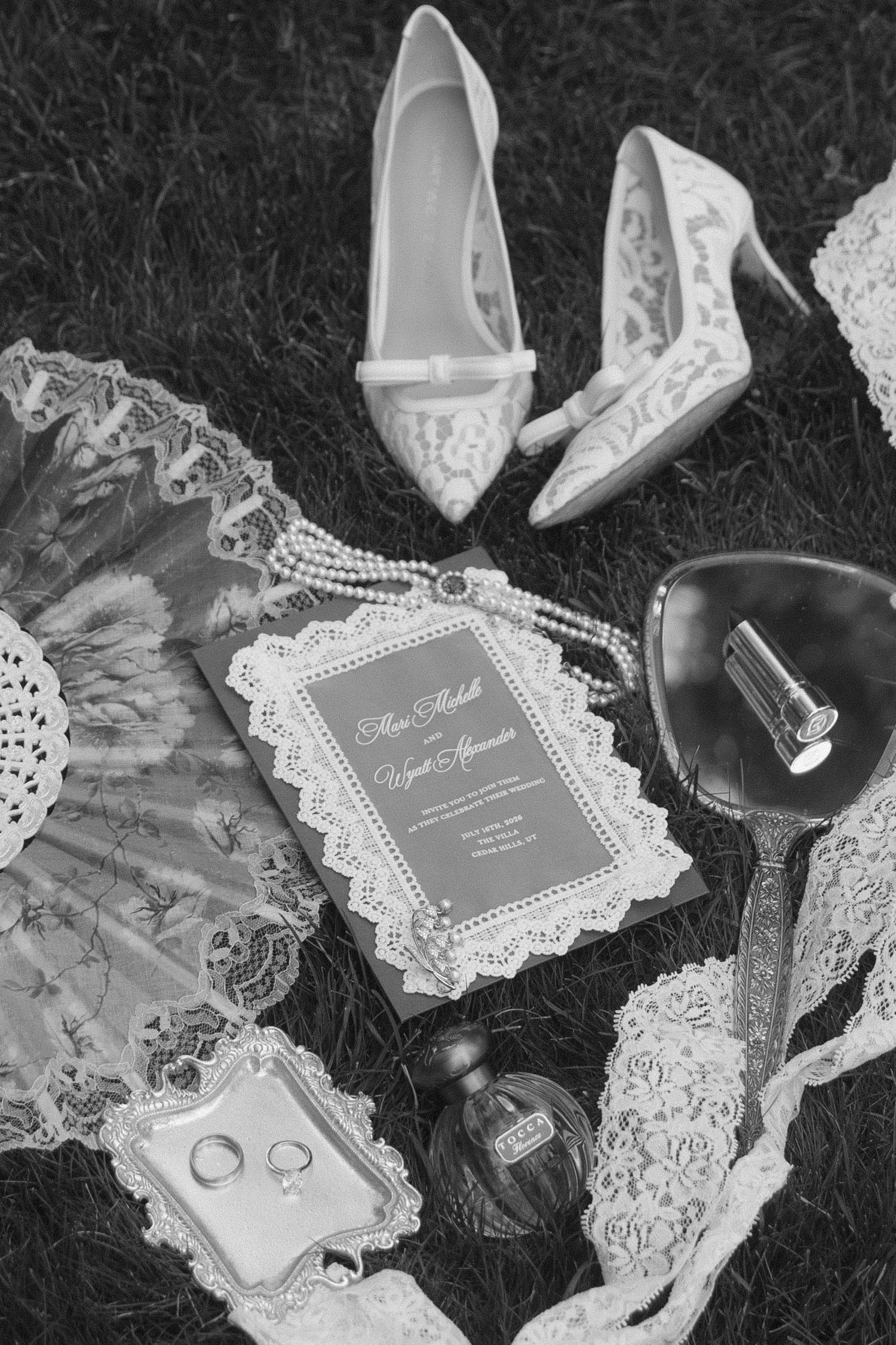 Black and white flat lay of bridal accessories including lace heels, pearl necklace, rings, perfume, hand mirror, and wedding invitation.