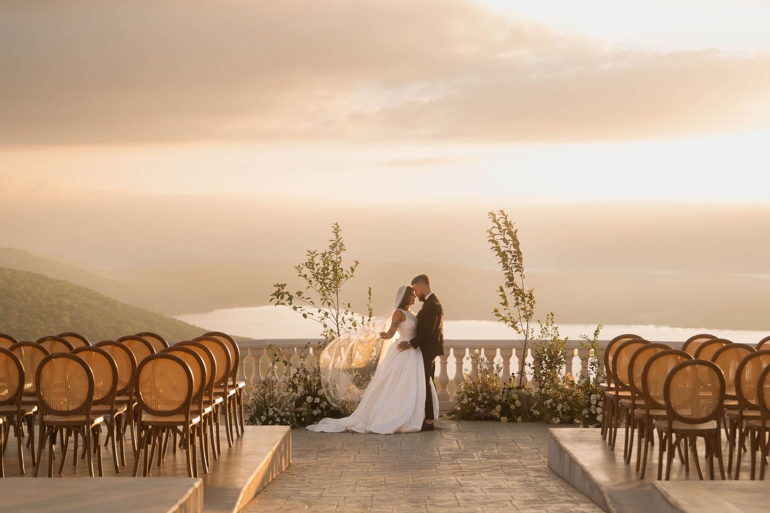 Bride and groom embrace at sunset outdoor wedding ceremony with rattan chairs and ocean view backdrop.