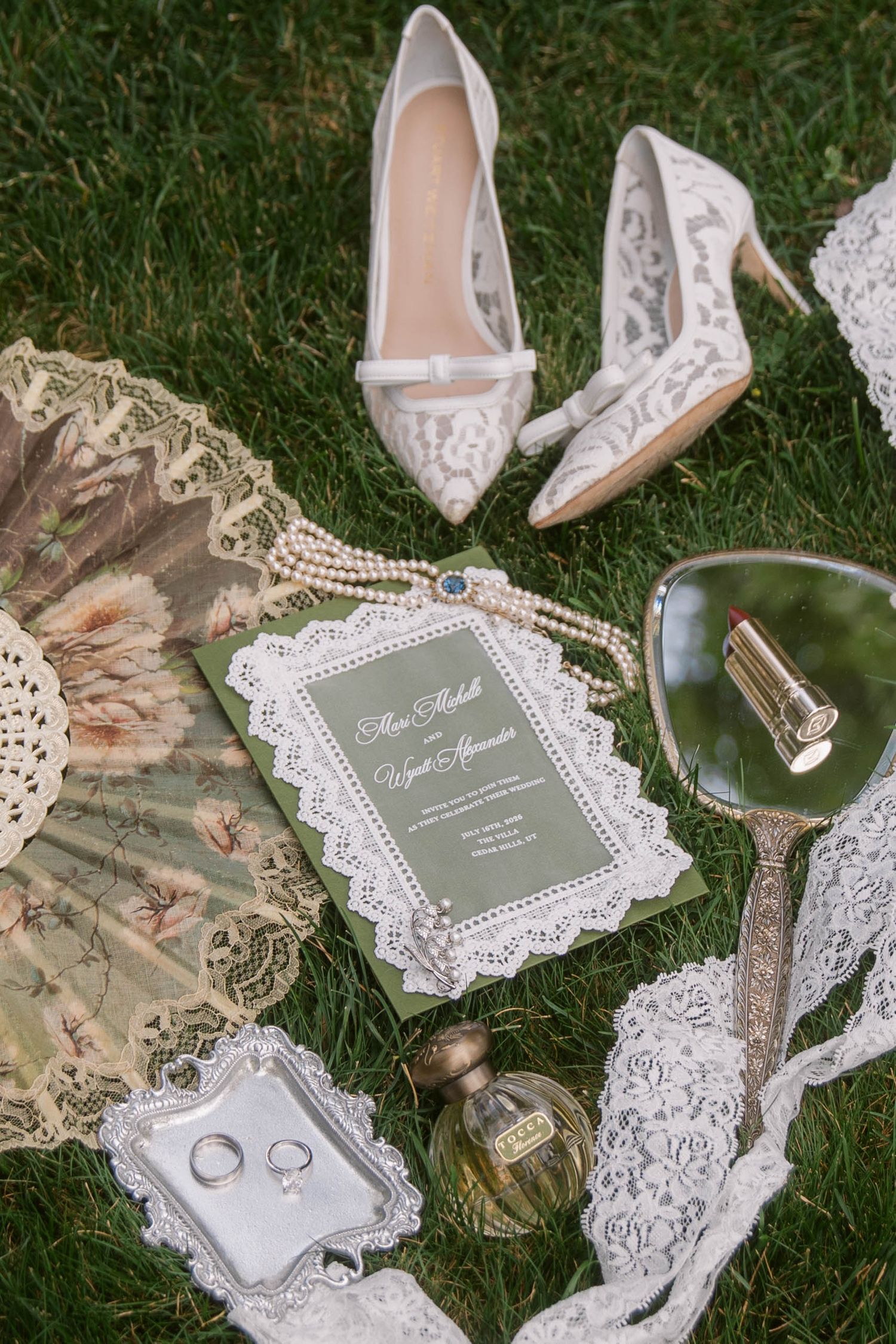 Elegant wedding flat lay with lace heels, pearl necklace, green invitation, vintage mirror, perfume, rings, and floral fan on grass.