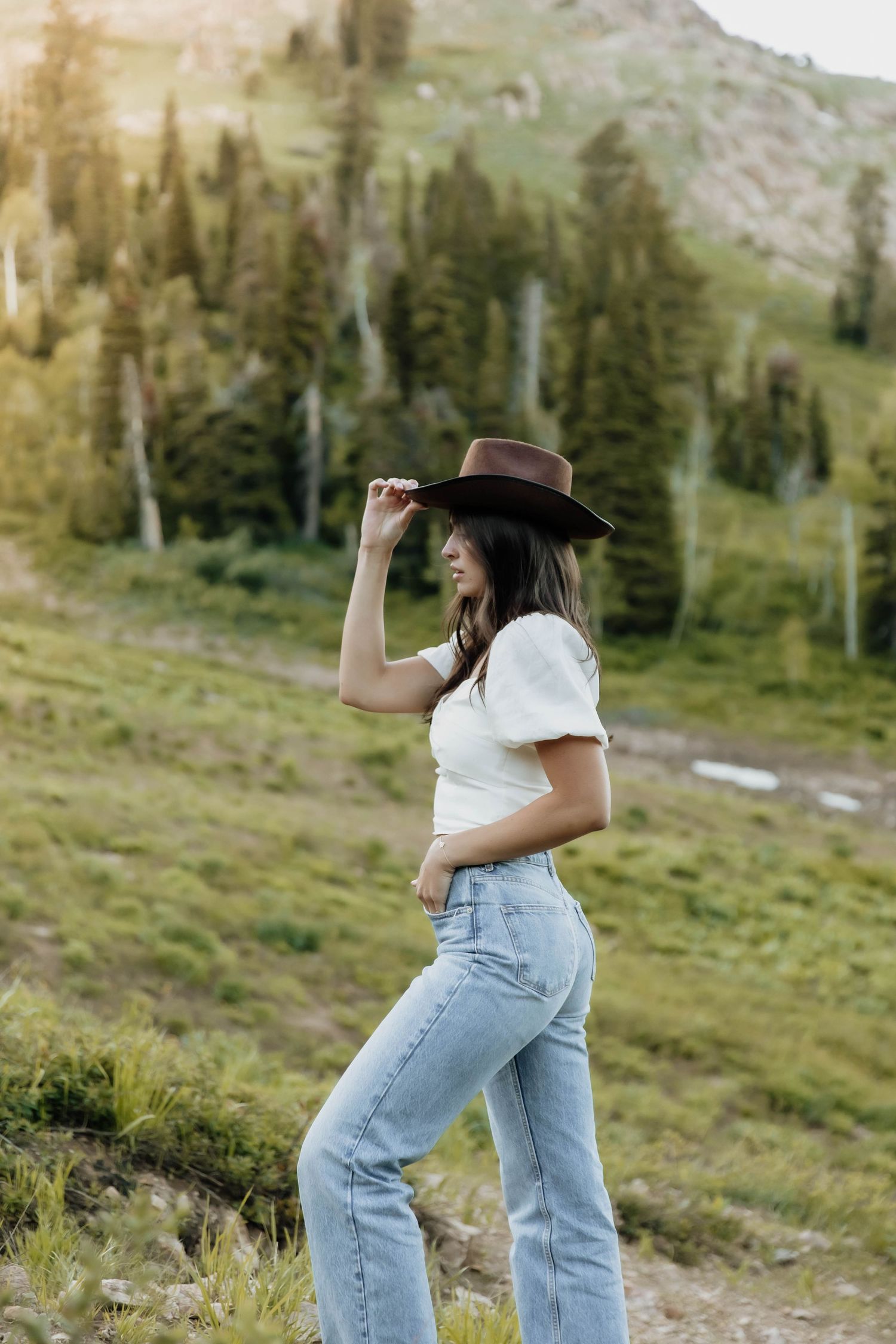 Brunette woman in white top and jeans tips brown cowboy hat while posing on mountain meadow trail.