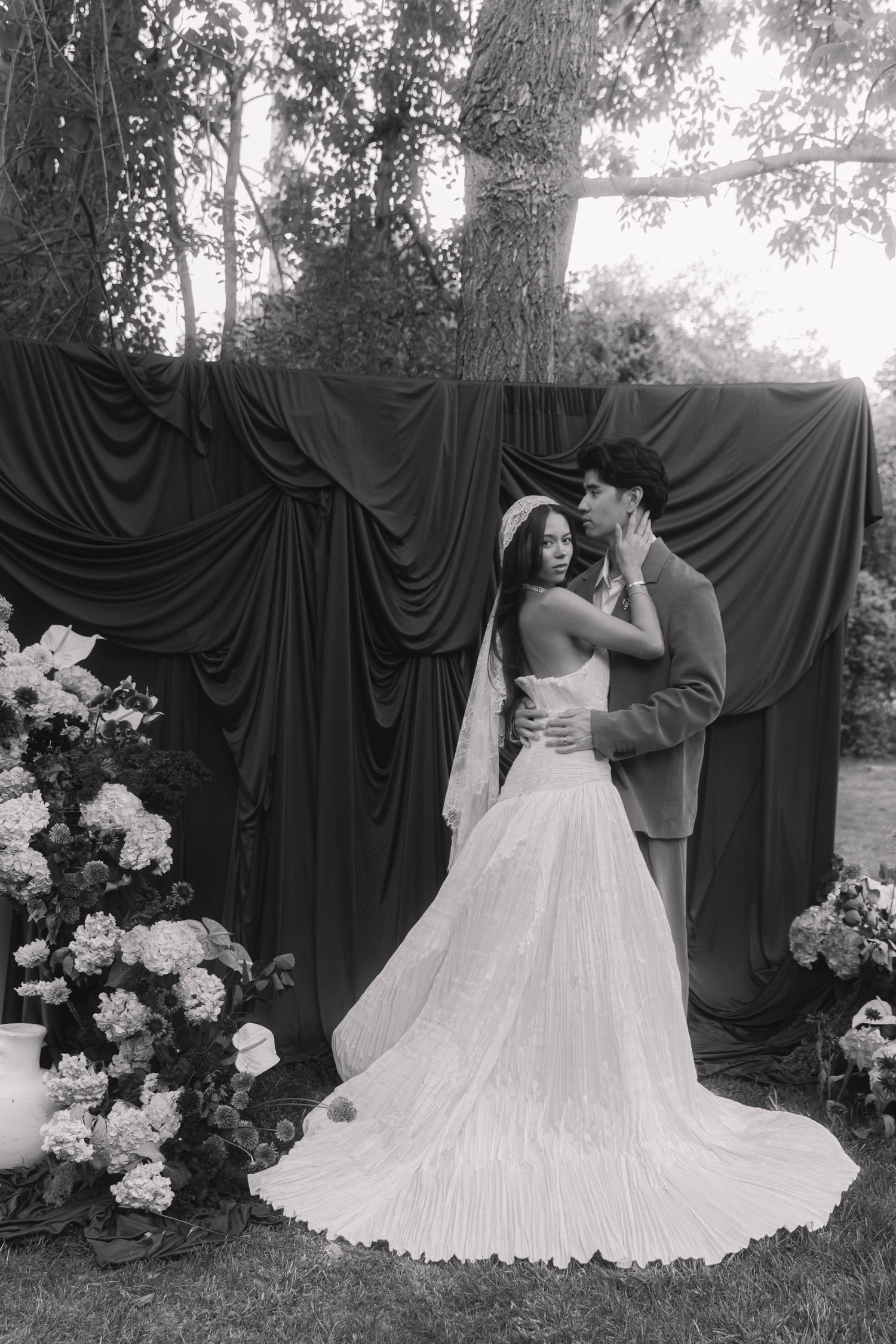 Black and white photo of a bride and groom embracing before draped fabric backdrop with floral arrangements outdoors.
