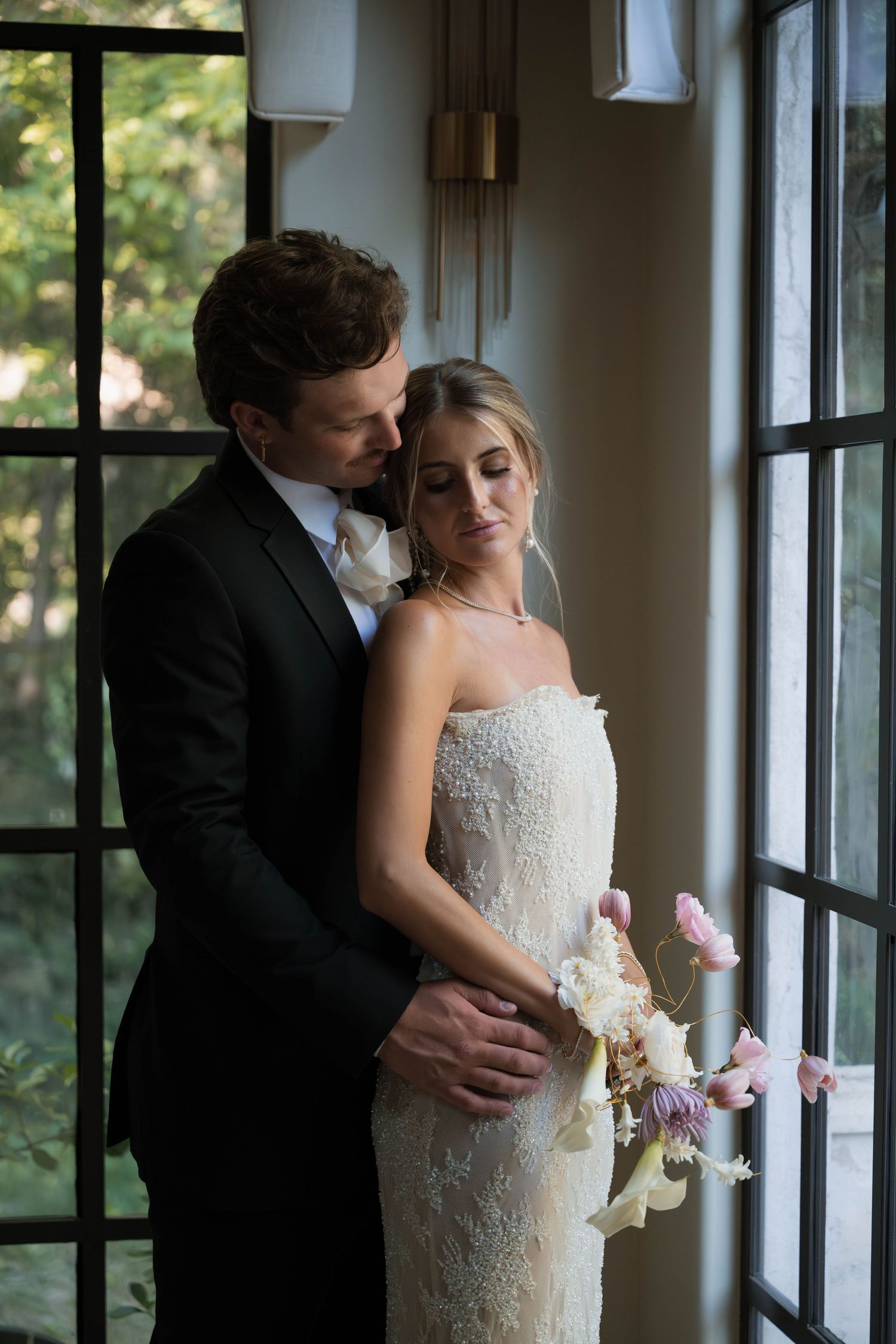 Groom embraces bride from behind as she holds a pink floral bouquet near large windows indoors.