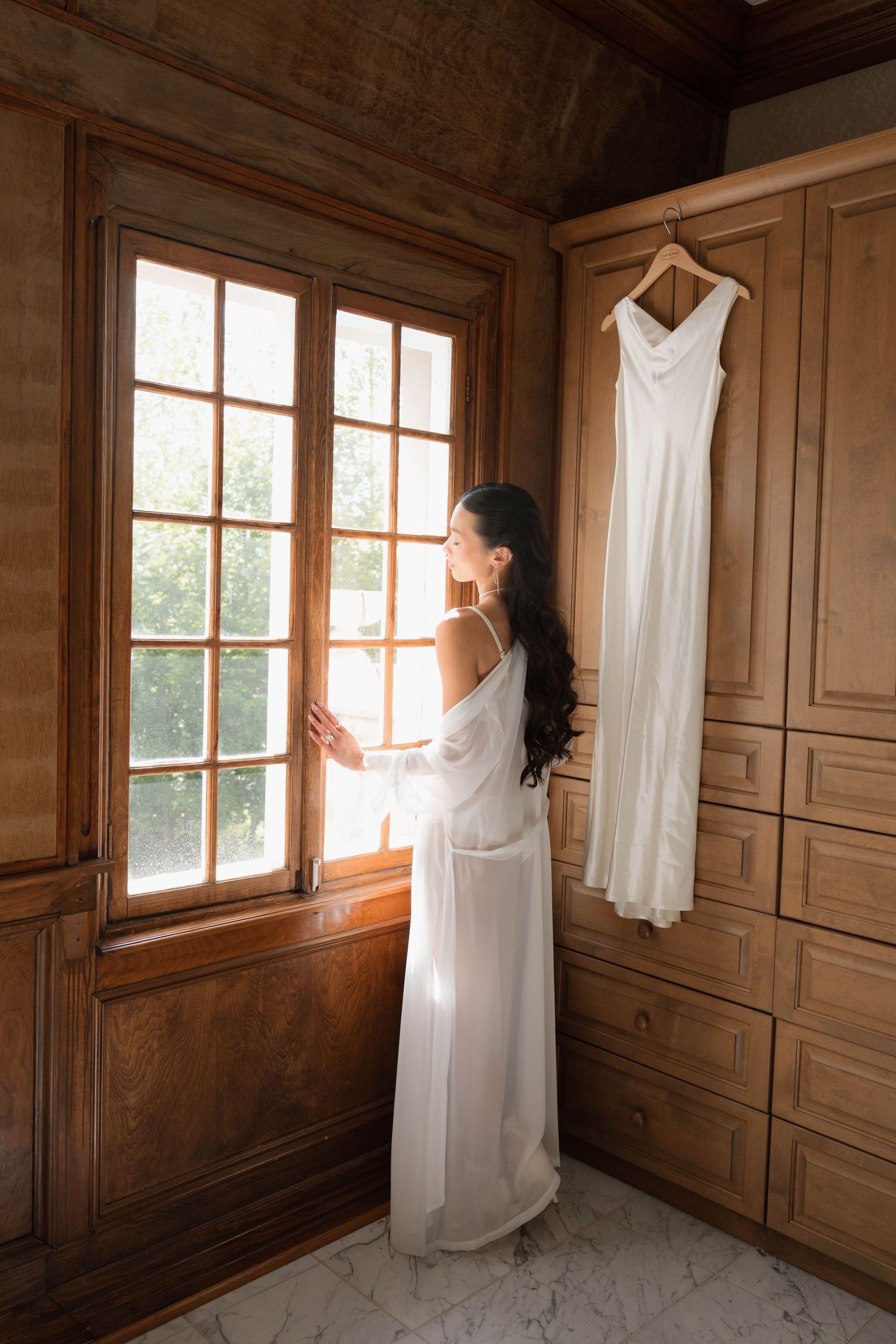 Bride in white robe gazes out sunlit window beside her hanging wedding dress in a wooden dressing room.