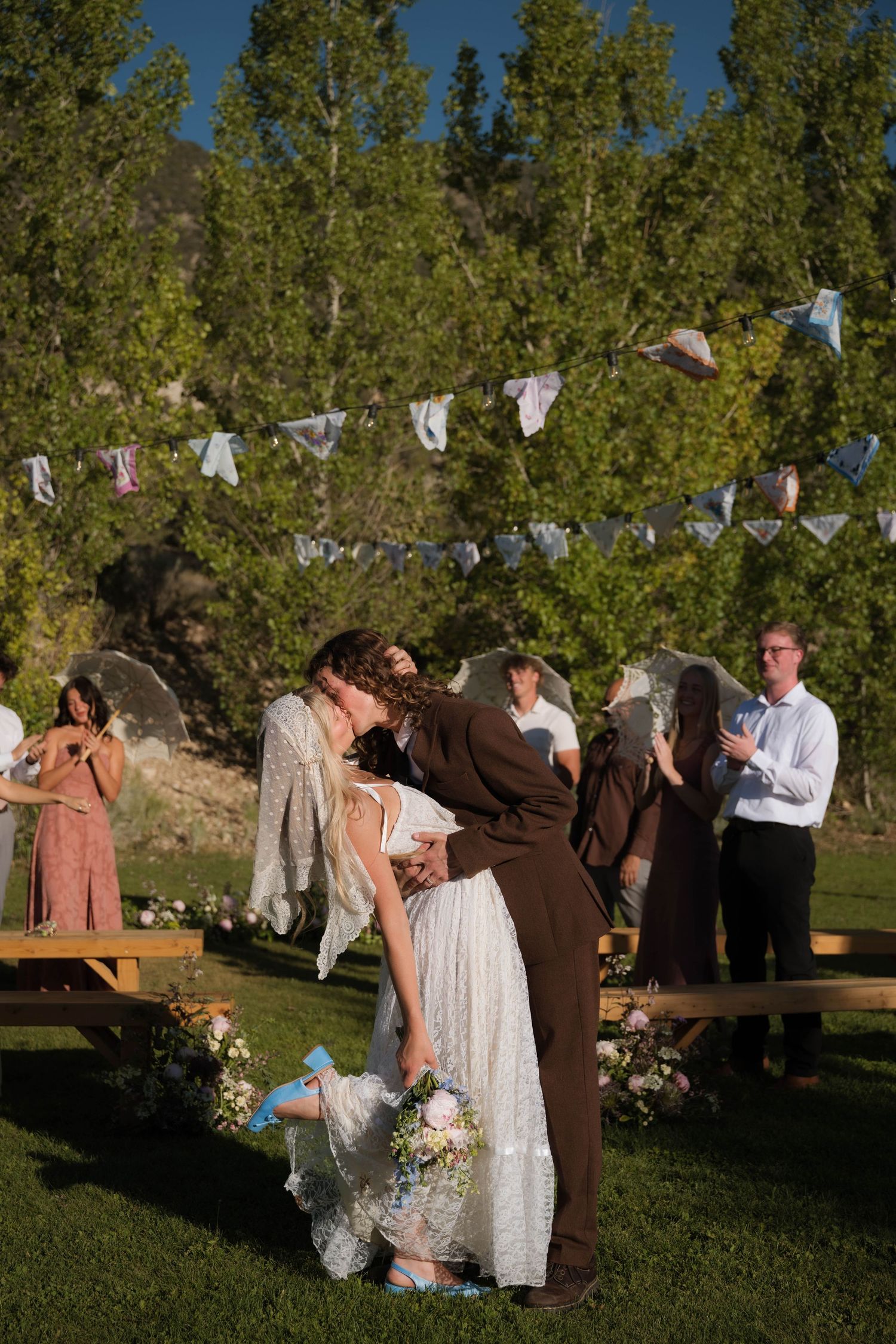 Couple sharing first kiss at outdoor wedding ceremony surrounded by guests and trees.