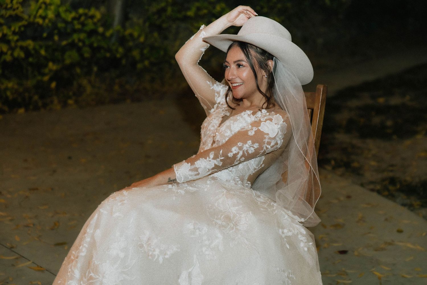 Smiling bride in a floral lace wedding gown and white cowboy hat seated outdoors at a rustic venue.