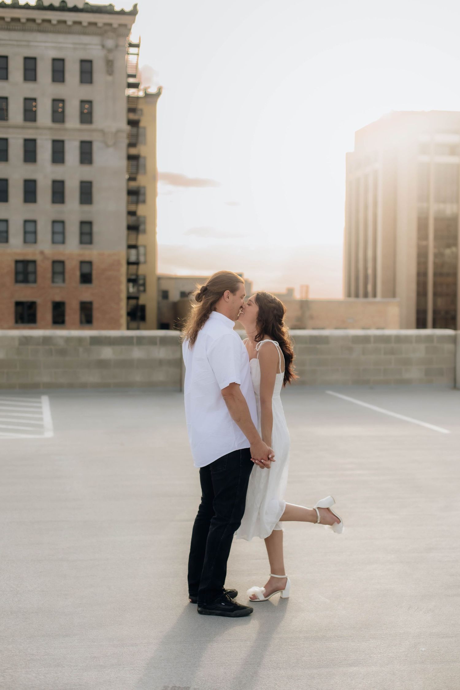 Couple shares a sunset kiss on urban rooftop parking deck surrounded by city buildings in golden light.