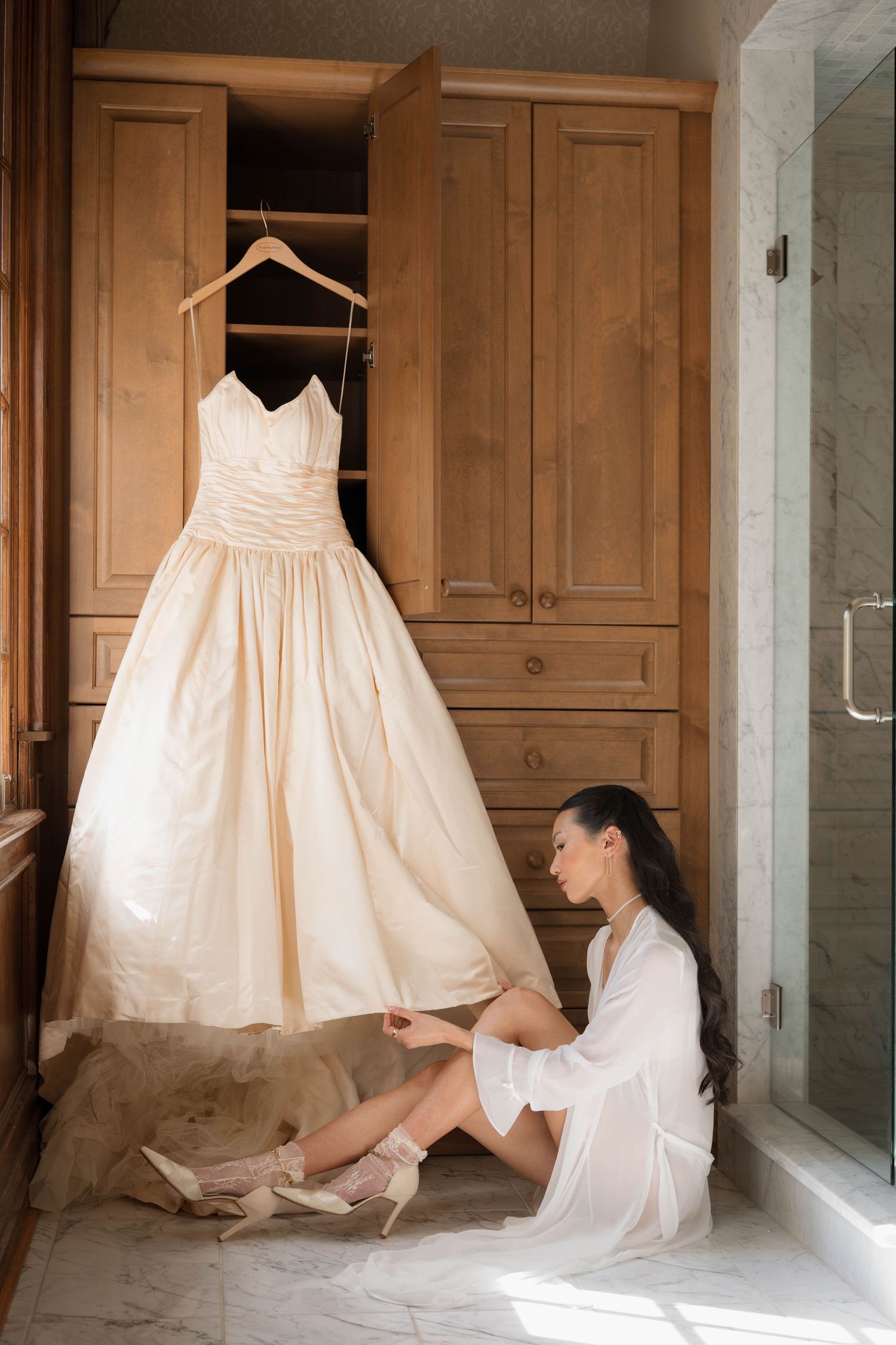 Bride in white robe sits on marble floor putting on heels beside her strapless ballgown wedding dress.