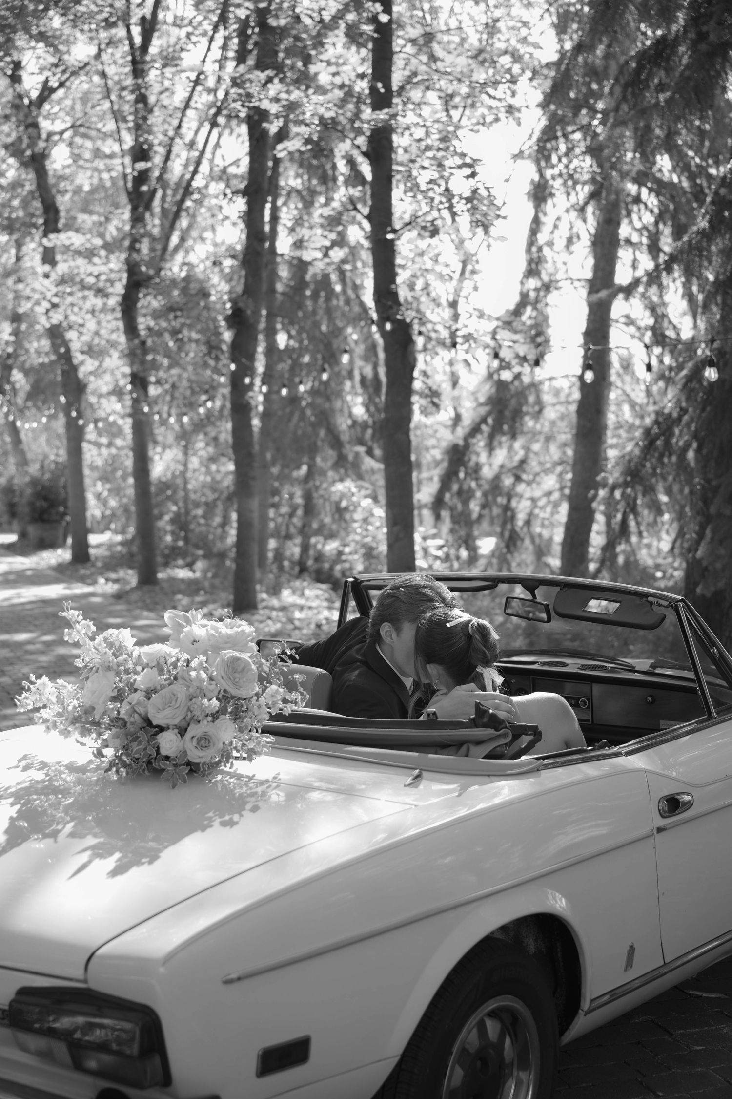 Black and white photo of a couple kissing in a vintage convertible with a bridal bouquet on the hood.