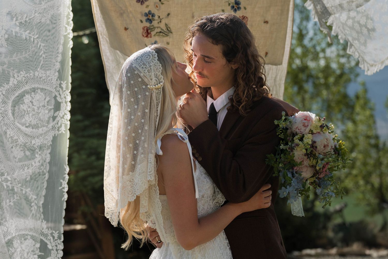 Two brides share a romantic moment at an outdoor wedding ceremony, one in a lace veil holding a bouquet.