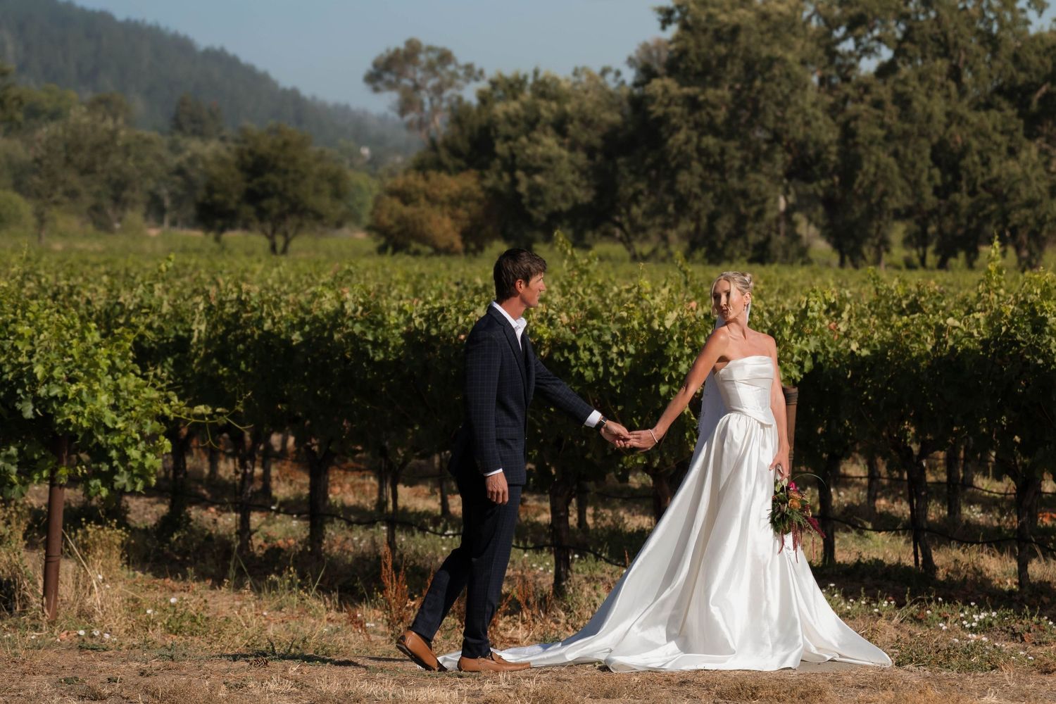 Bride and groom holding hands walking through a sunlit vineyard, bride in strapless gown, groom in navy suit.