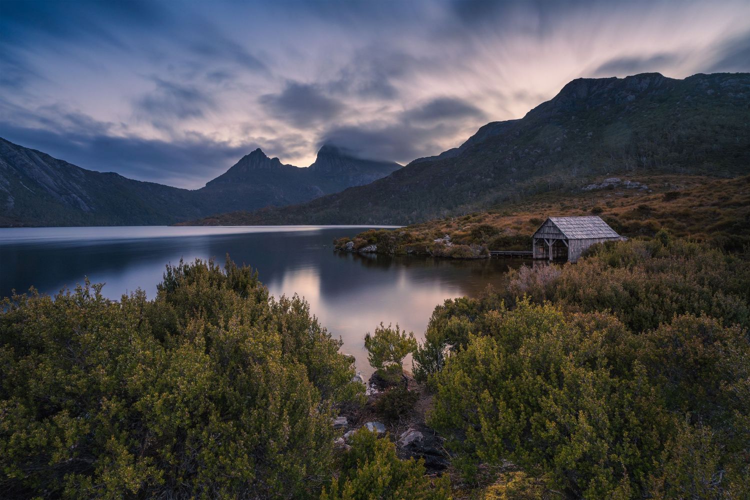 Rustic boathouse on a calm lake at dusk with dramatic cloudy mountains in the background.