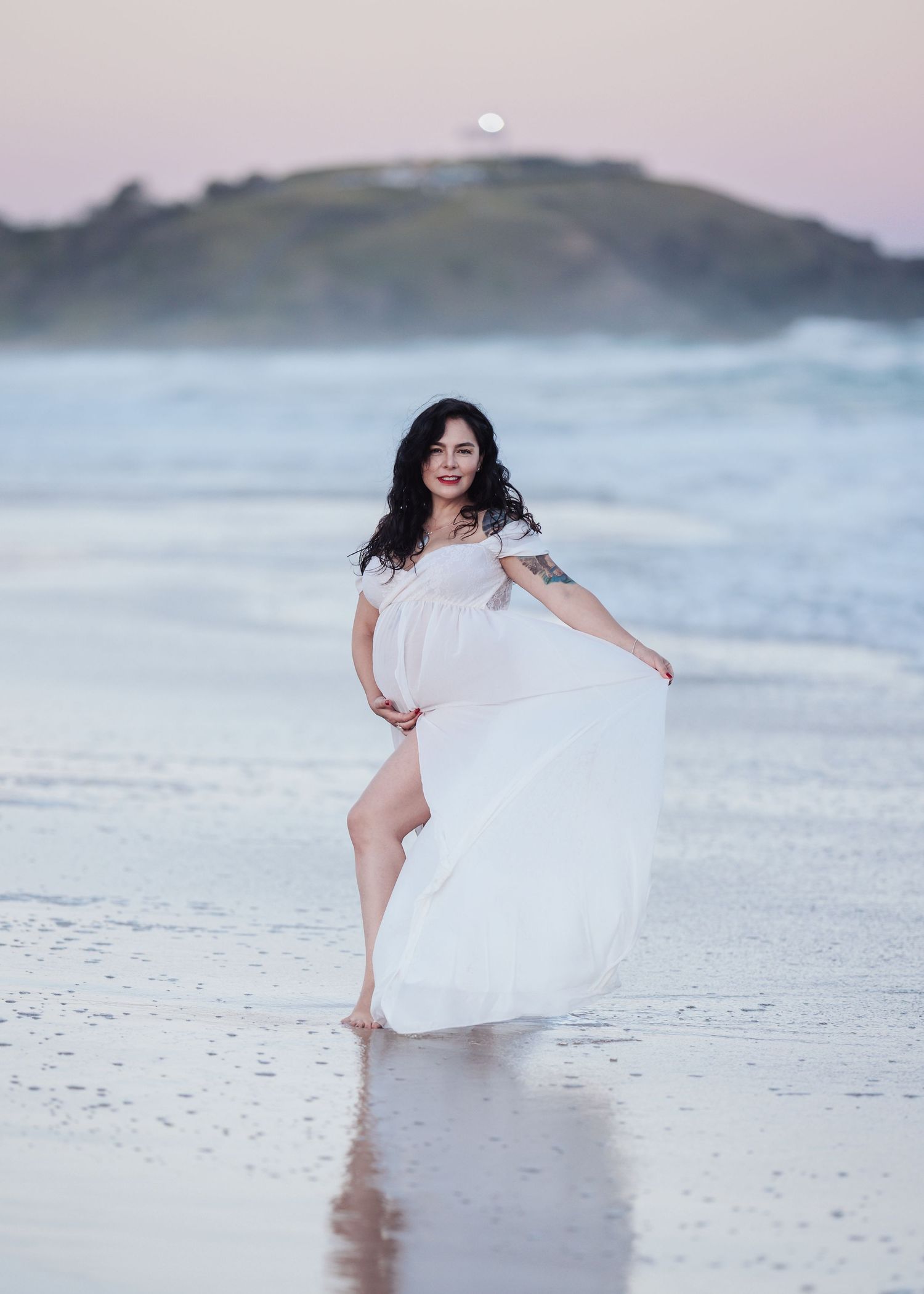 Woman in white flowing dress poses gracefully on beach at sunset with ocean waves behind her.