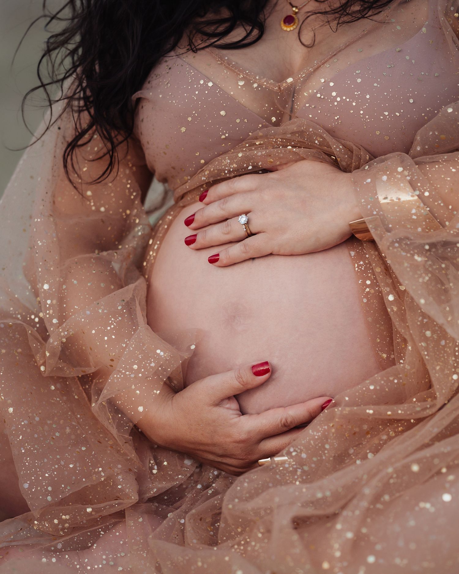 Close-up of pregnant belly with red nails and diamond ring in a sparkly beige tulle dress.