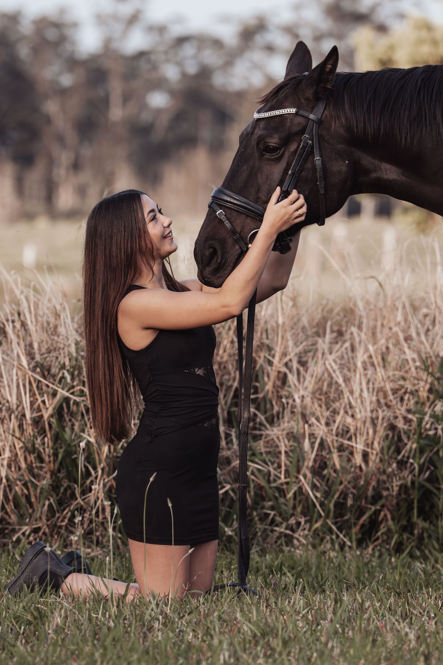 Young woman in black dress kneeling on grass, tenderly petting a dark horse's face in a rural field setting.