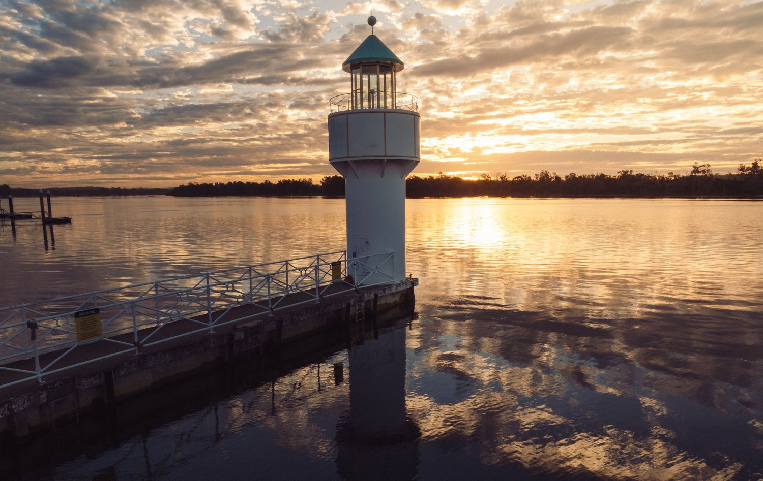 A white lighthouse on a dock reflects in calm water during a stunning golden sunset with dramatic cloudy skies.