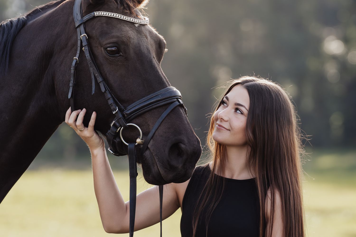 Smiling young woman with long brown hair gently holding a dark horse's bridle outdoors in soft light.