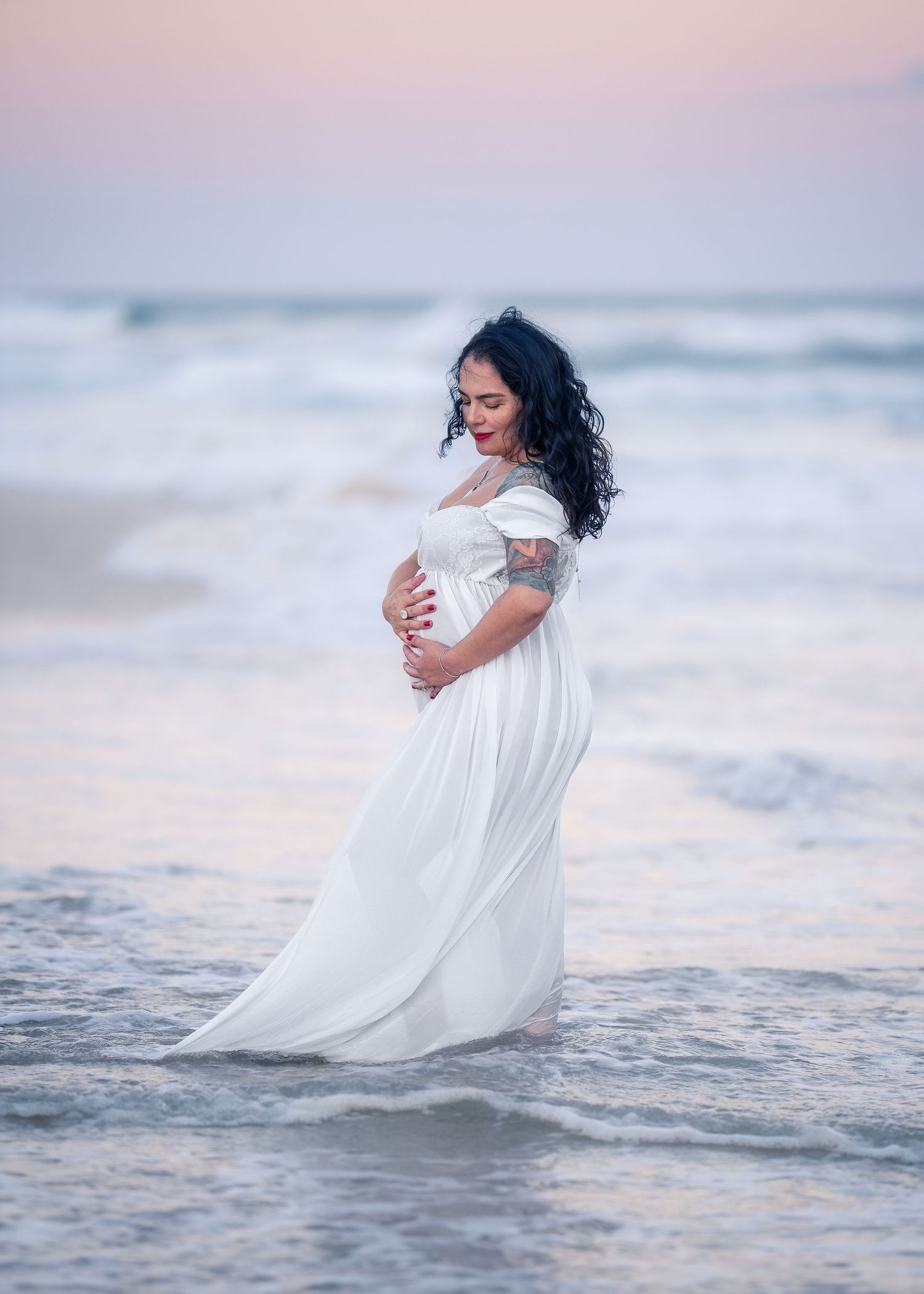 Beautiful pregnant woman in flowing white dress smiles while cradling bump in gentle ocean waves.
