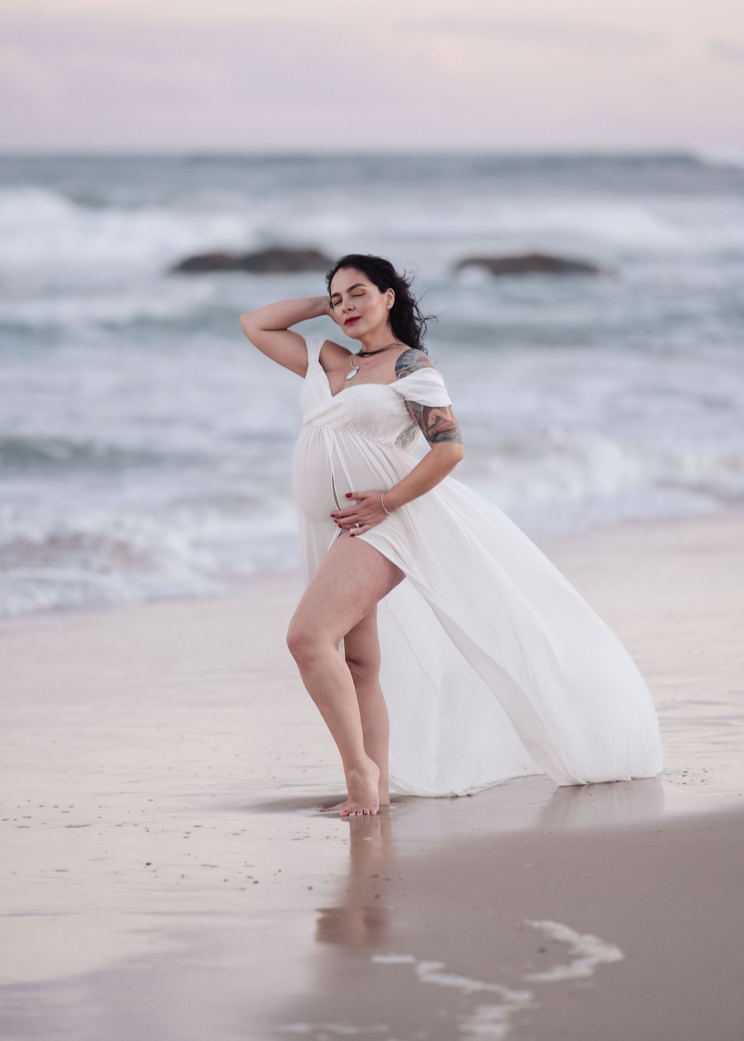 Pregnant woman in white chiffon dress cradles baby bump while standing barefoot on sandy beach.