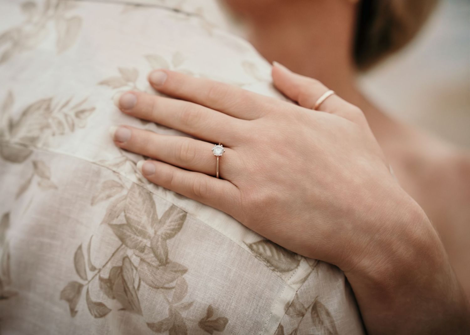Close-up of a woman's hand with a diamond engagement ring resting on a man's floral shirt shoulder.