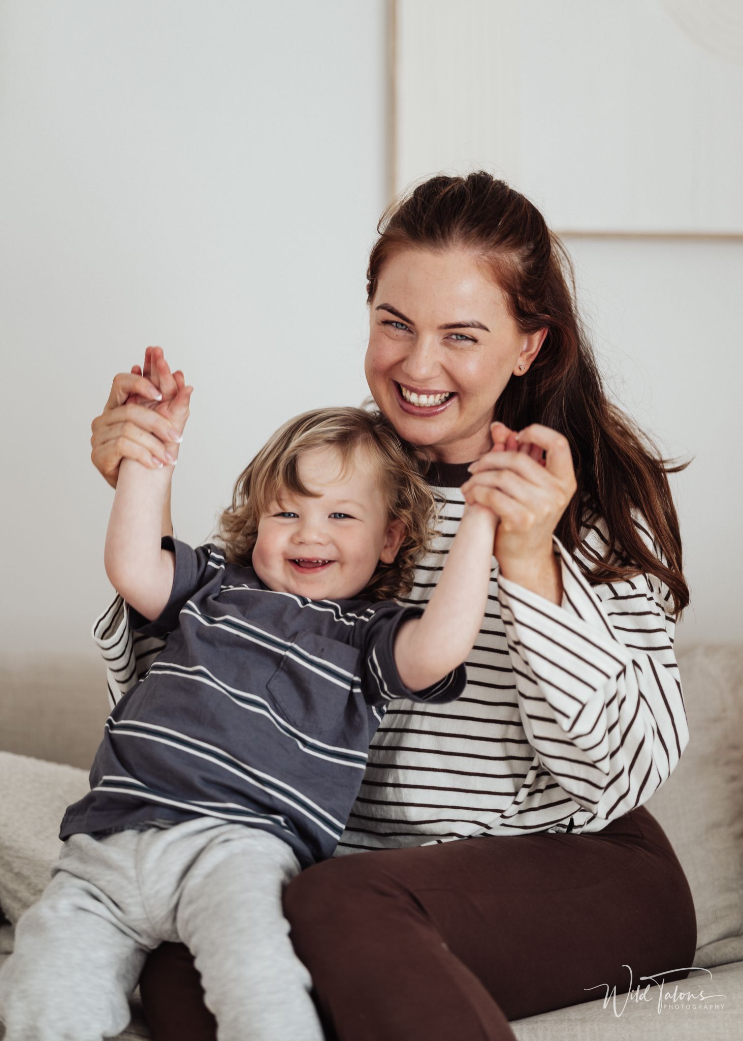A laughing woman in a striped top sits with a joyful toddler raising their arms on a white couch.