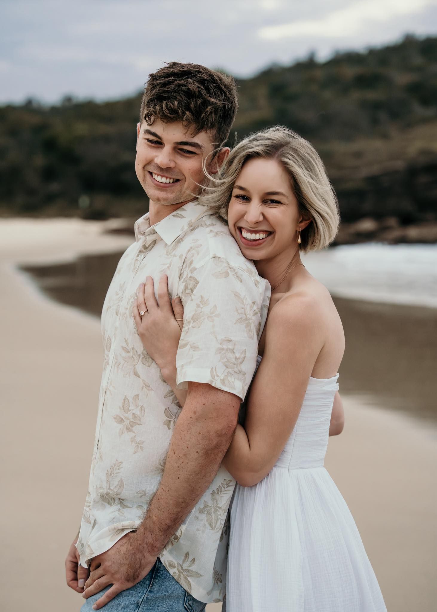 Smiling couple embracing on a sandy beach, woman in white dress hugging man in floral shirt.