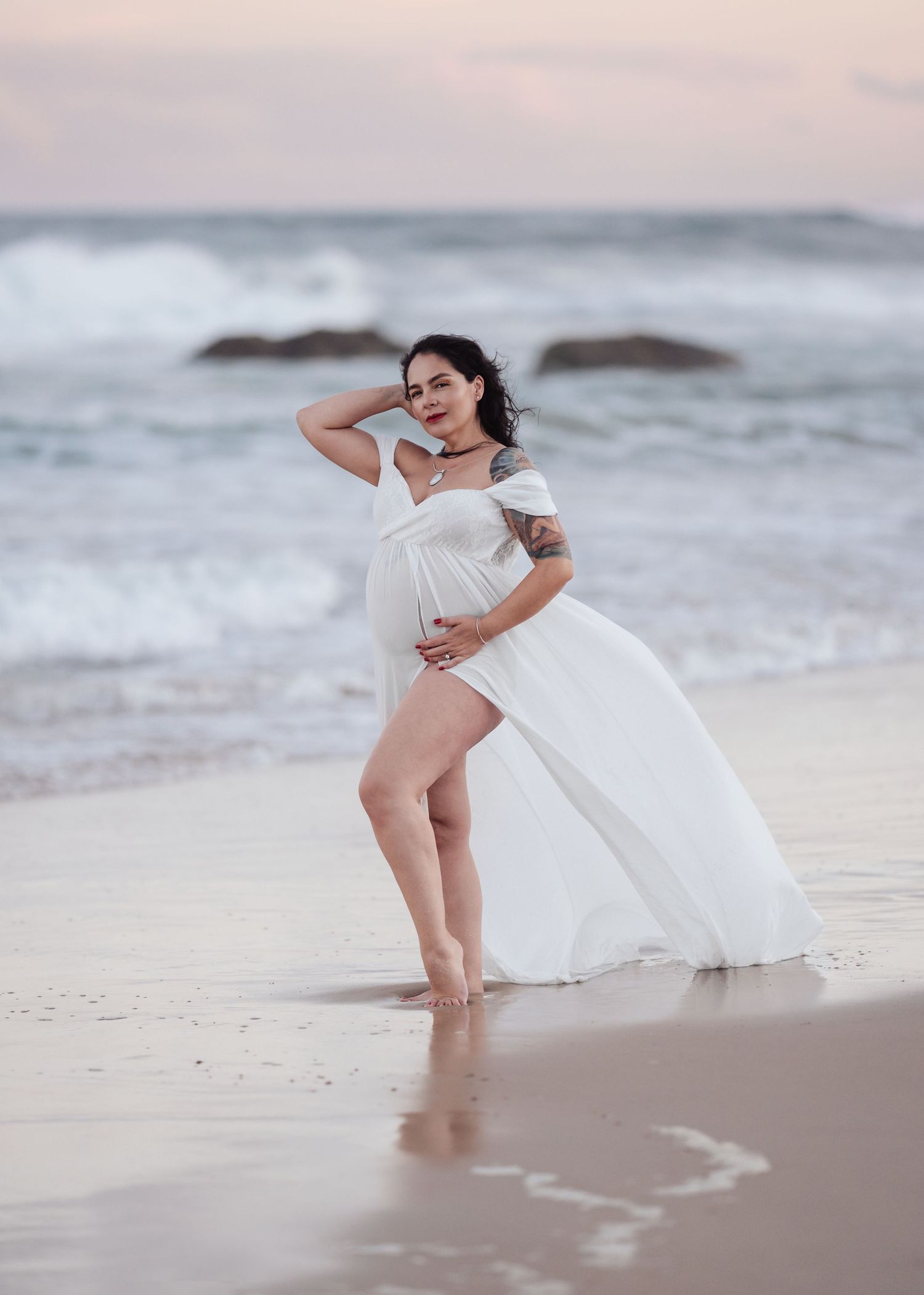 Maternity photo of woman in white off-shoulder dress posing on sandy beach at dusk.