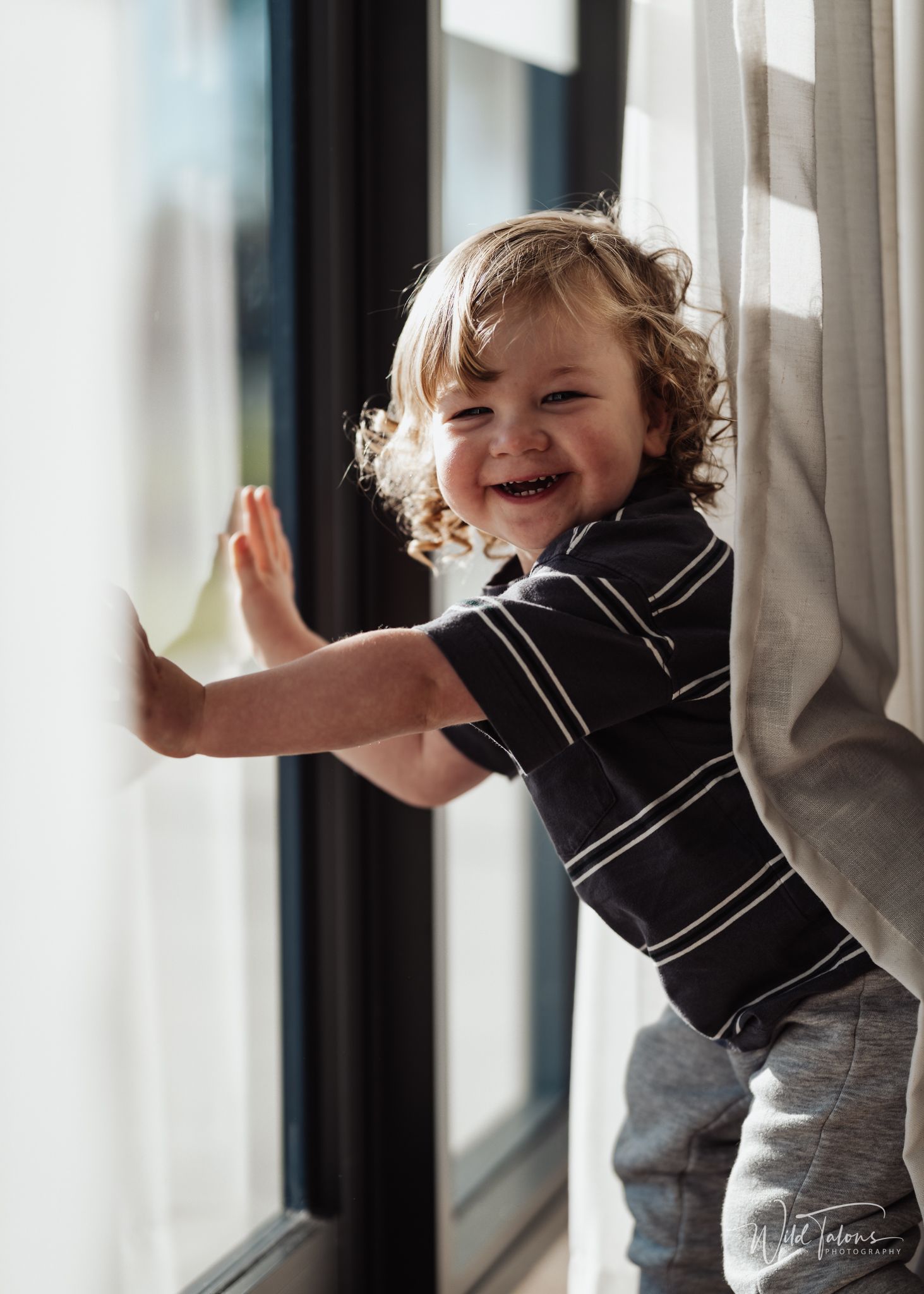 Curly-haired toddler boy laughing joyfully while leaning against a sunlit window indoors.