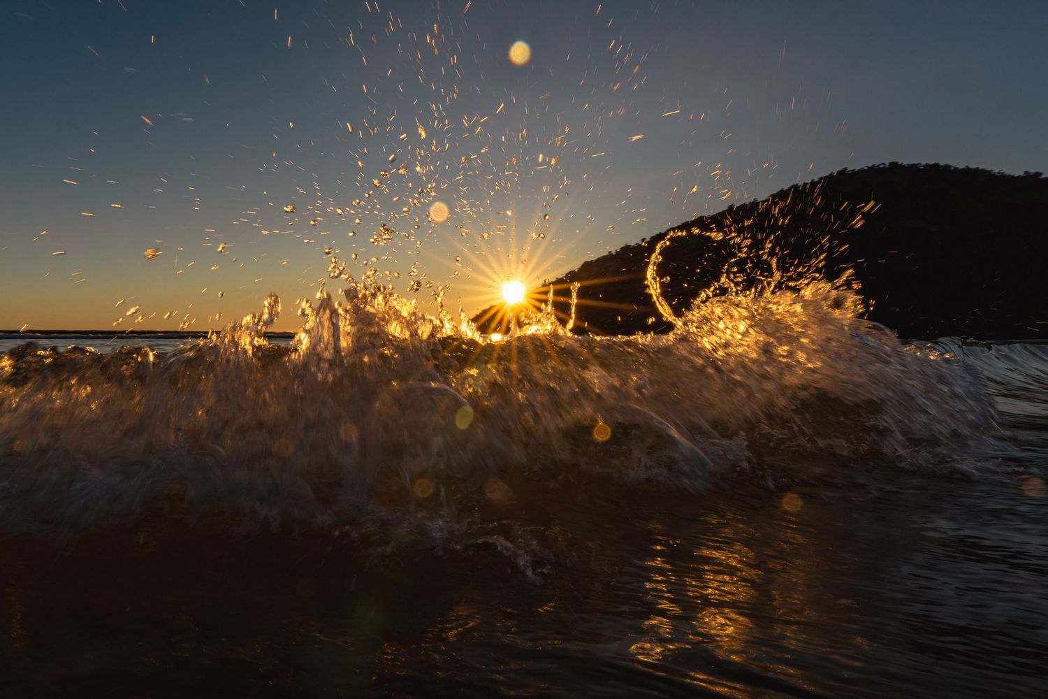 Golden sunburst bursts through a crashing ocean wave at sunset, creating a stunning splash of light and water.