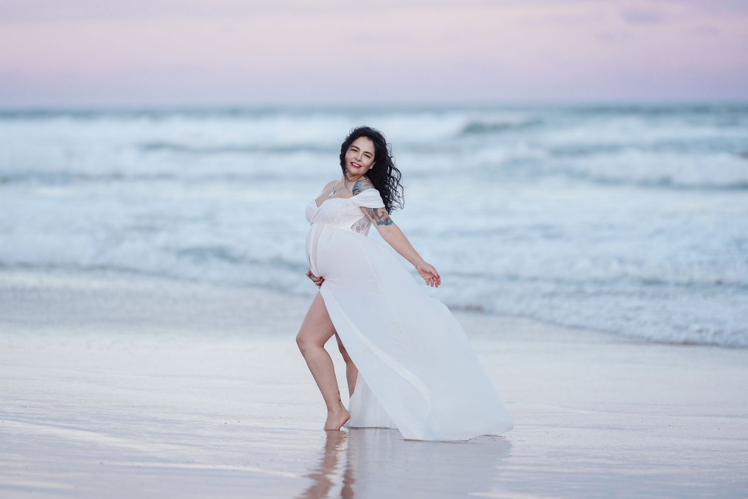 Pregnant woman in white flowing dress twirling on beach shoreline with waves in background.