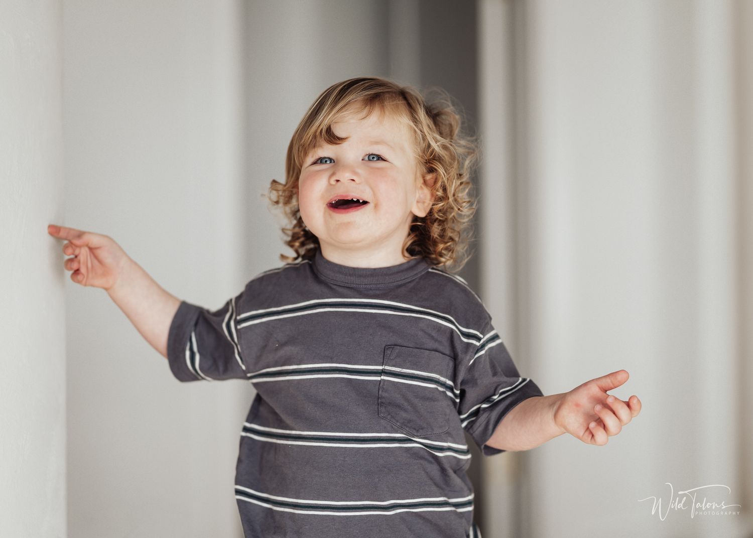 Joyful toddler with curly hair wearing a striped gray shirt, arms outstretched, smiling and laughing indoors.