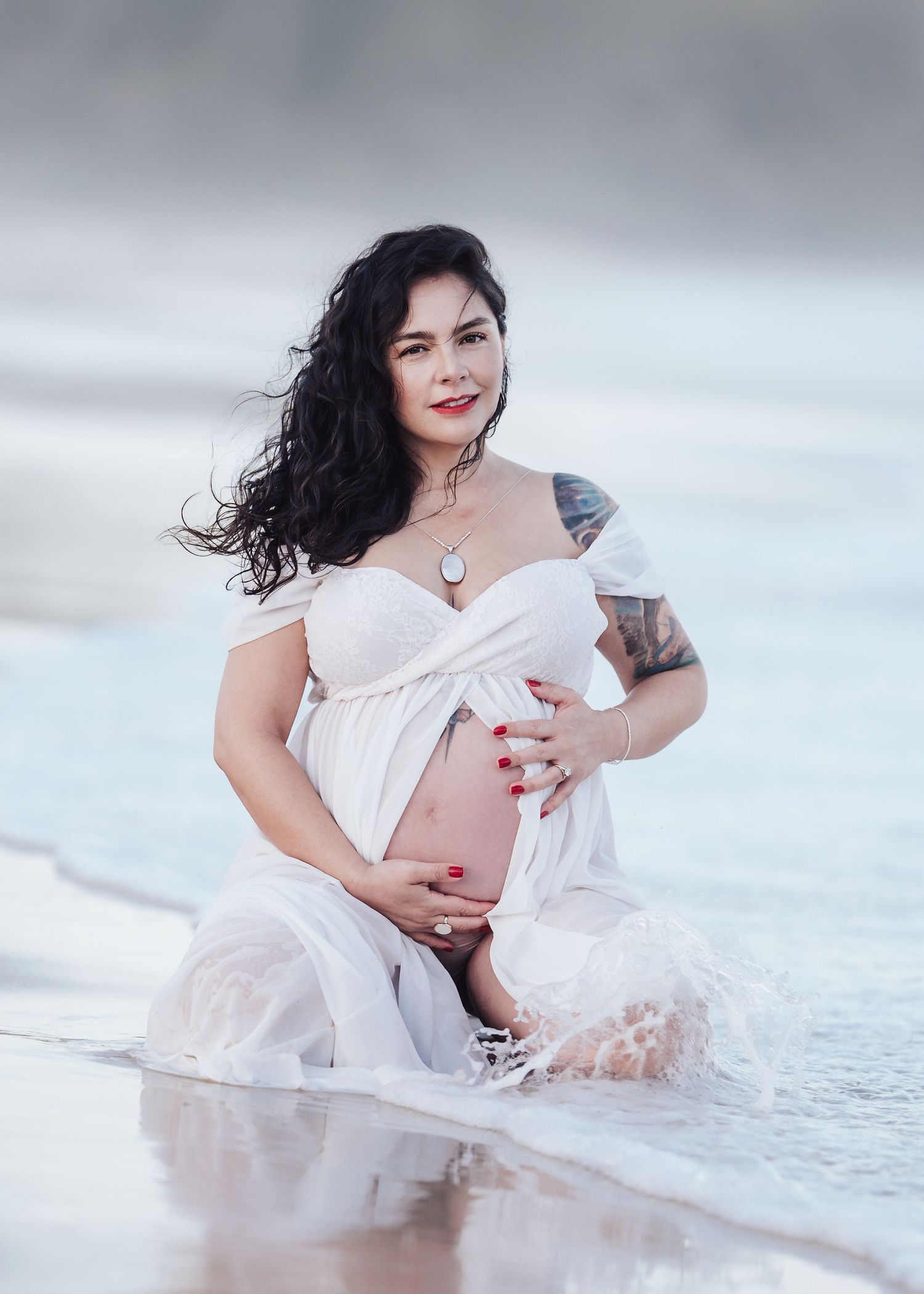Smiling pregnant woman in white dress kneeling in ocean waves during beach maternity session.