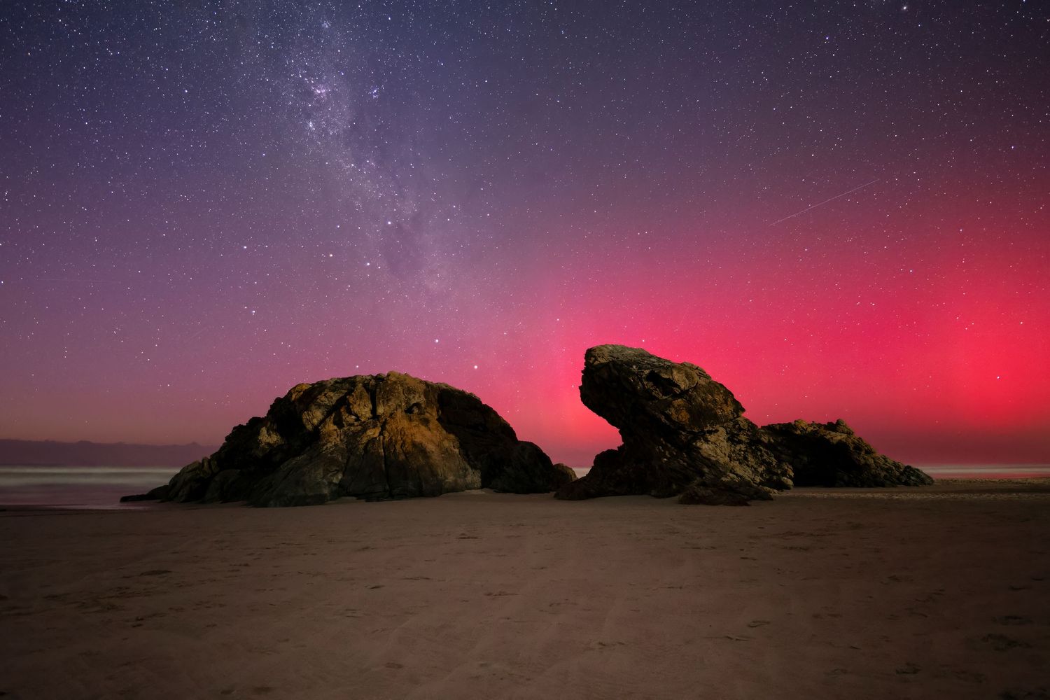 Rocky beach at night with vibrant red aurora borealis and Milky Way stars illuminating the dramatic coastal landscape.