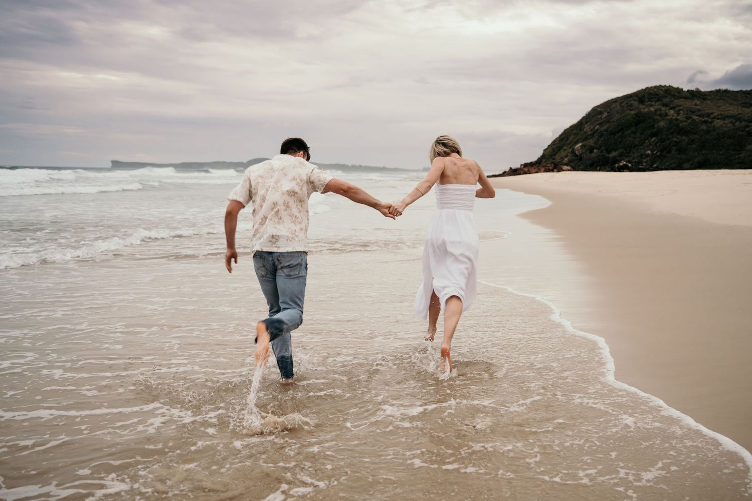 Couple running hand in hand through ocean waves on a sandy beach with green hills behind.