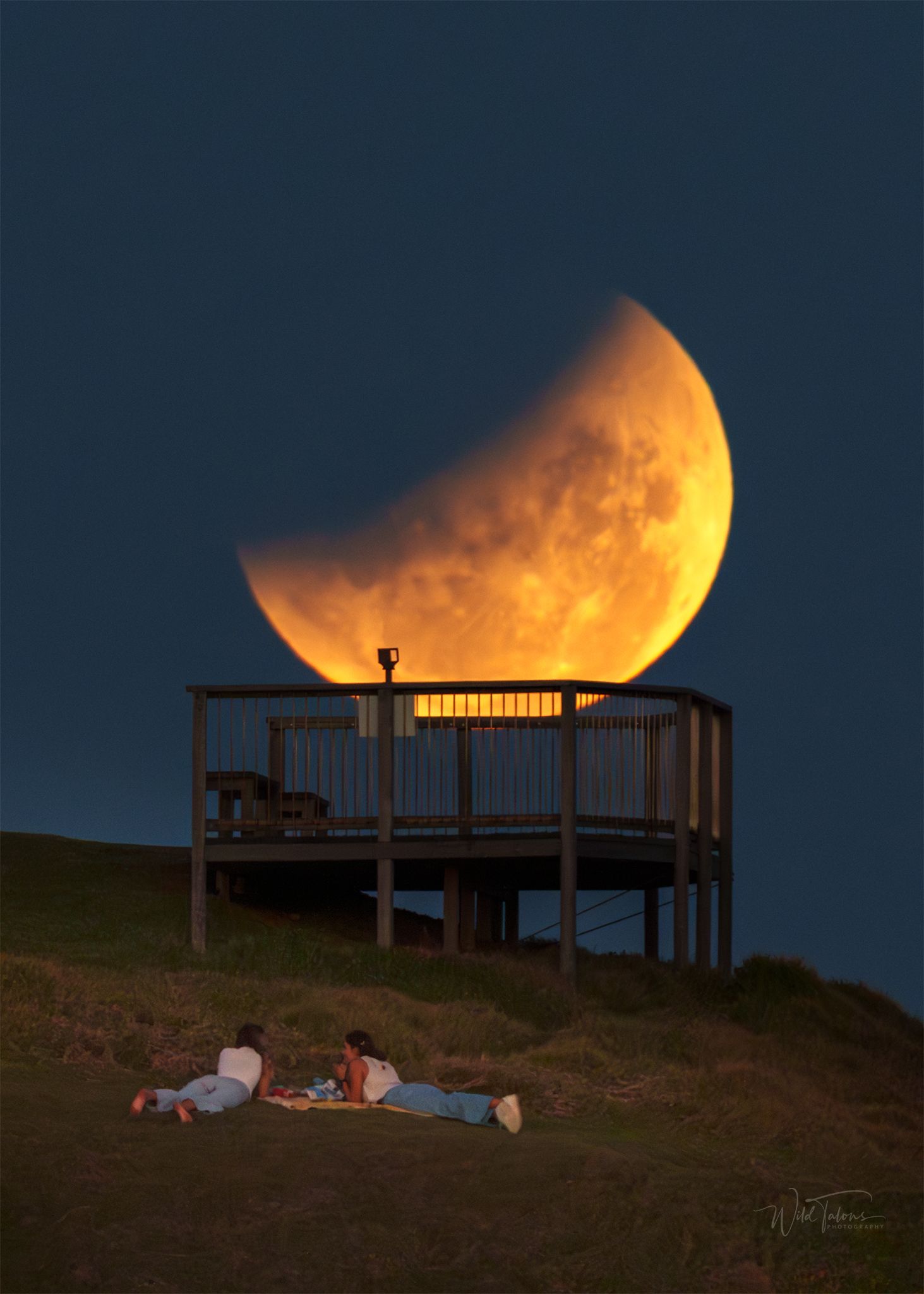 Two people lying on a hillside watching a dramatic crescent moon lunar eclipse rising behind a wooden observation deck at twilight.