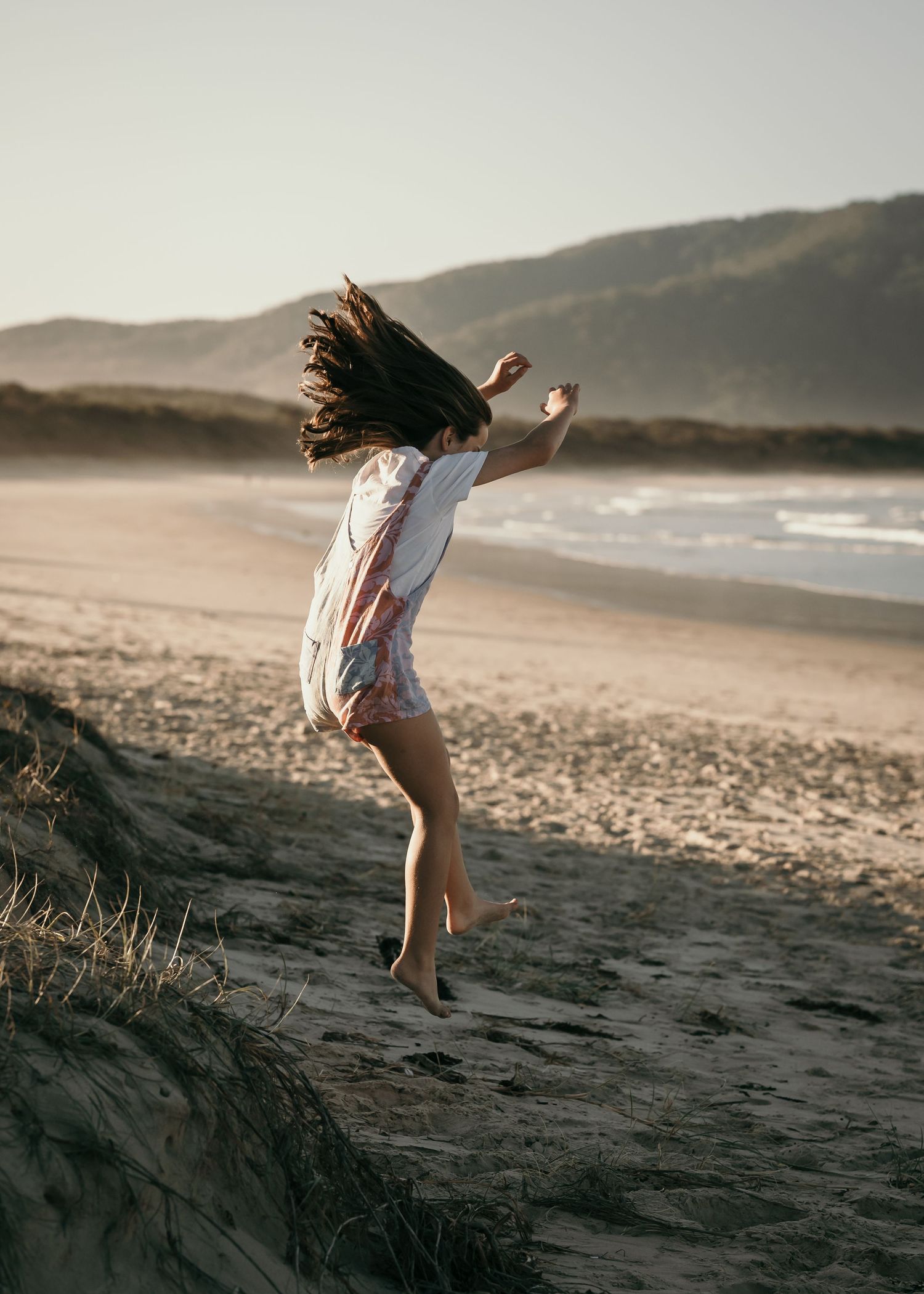 A girl with long dark hair jumps joyfully on a sandy beach at sunset with mountains in the background.