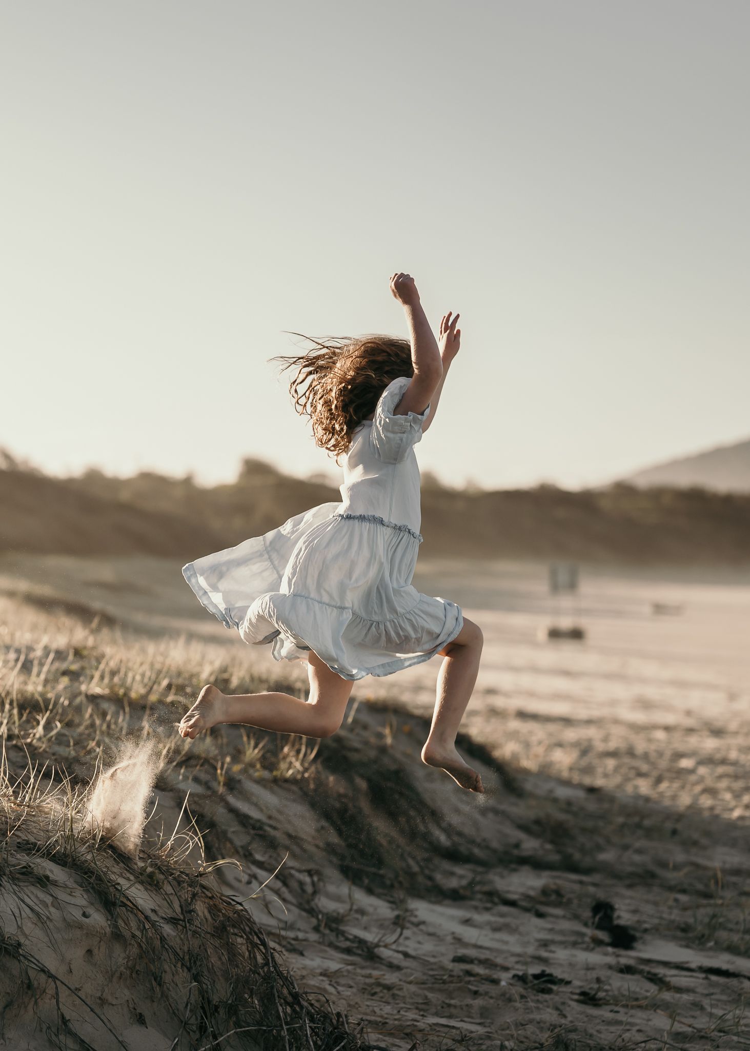A young girl in a white dress leaps joyfully on sandy dunes at golden hour near the beach.