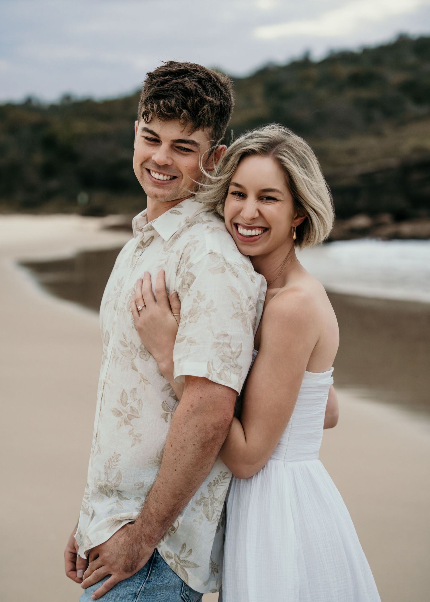 Laughing young couple embracing on sandy beach with ocean backdrop at dusk.