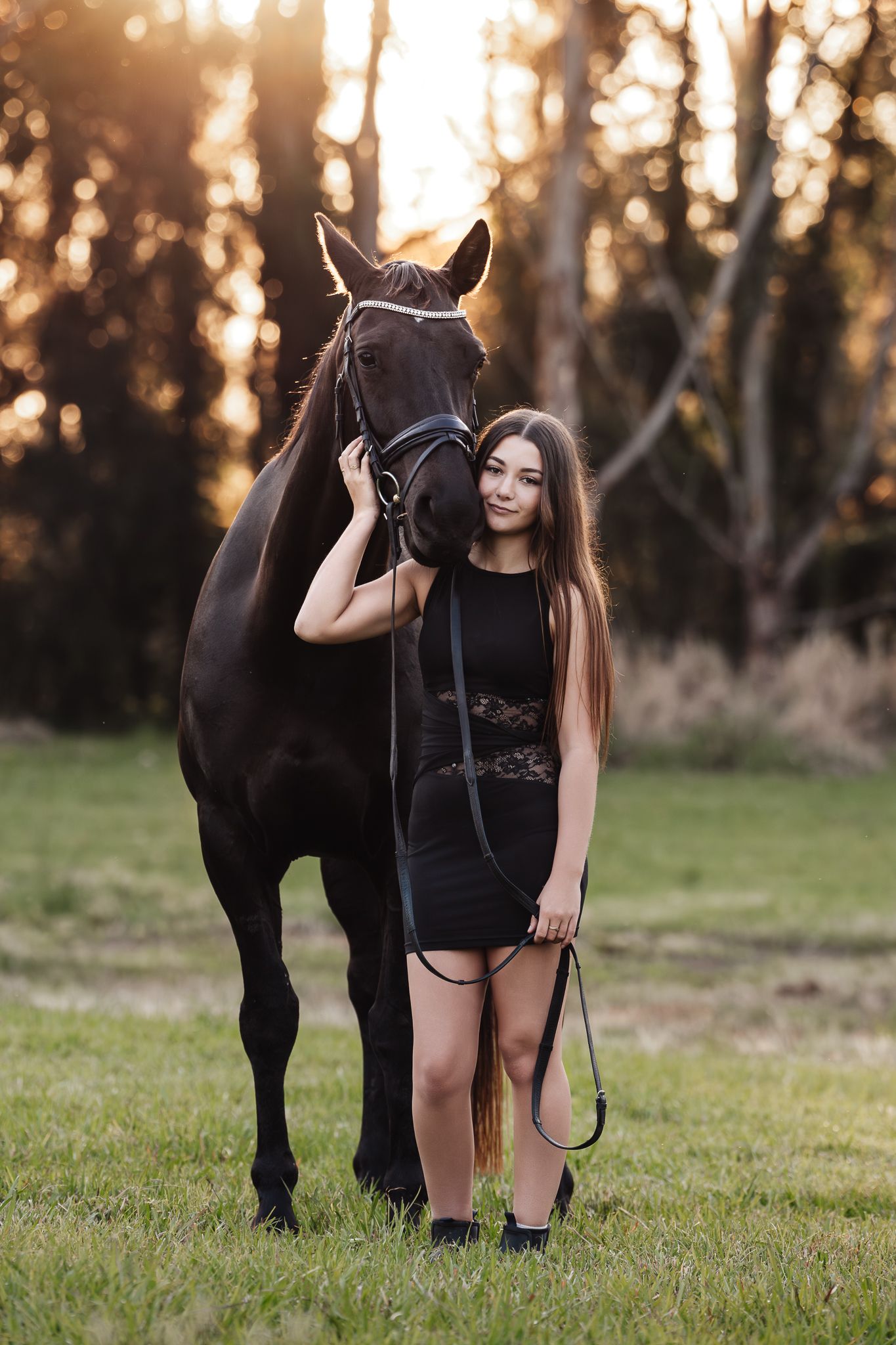 Young woman in black dress standing beside a dark horse on green grass during a warm golden sunset.