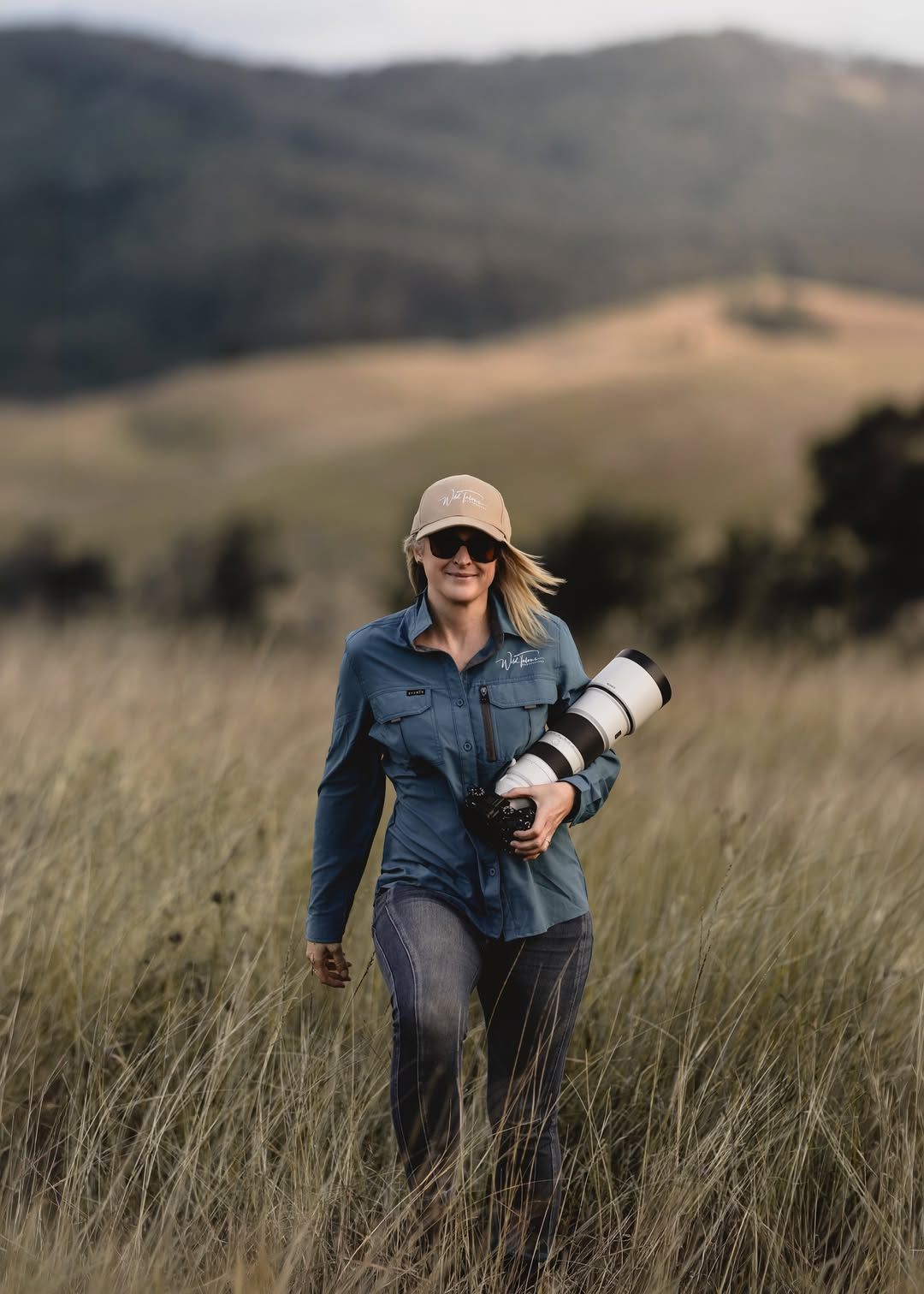 Woman photographer walking through tall grass fields with a telephoto lens, mountains in the background.