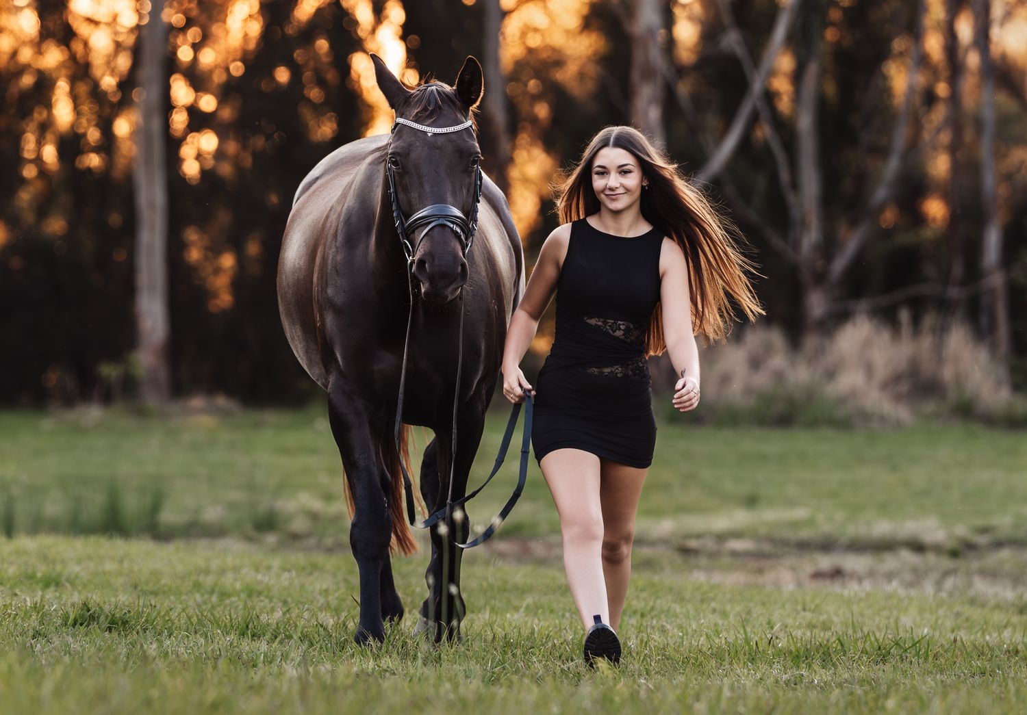 Young woman in black dress walks dark horse through golden sunset field.