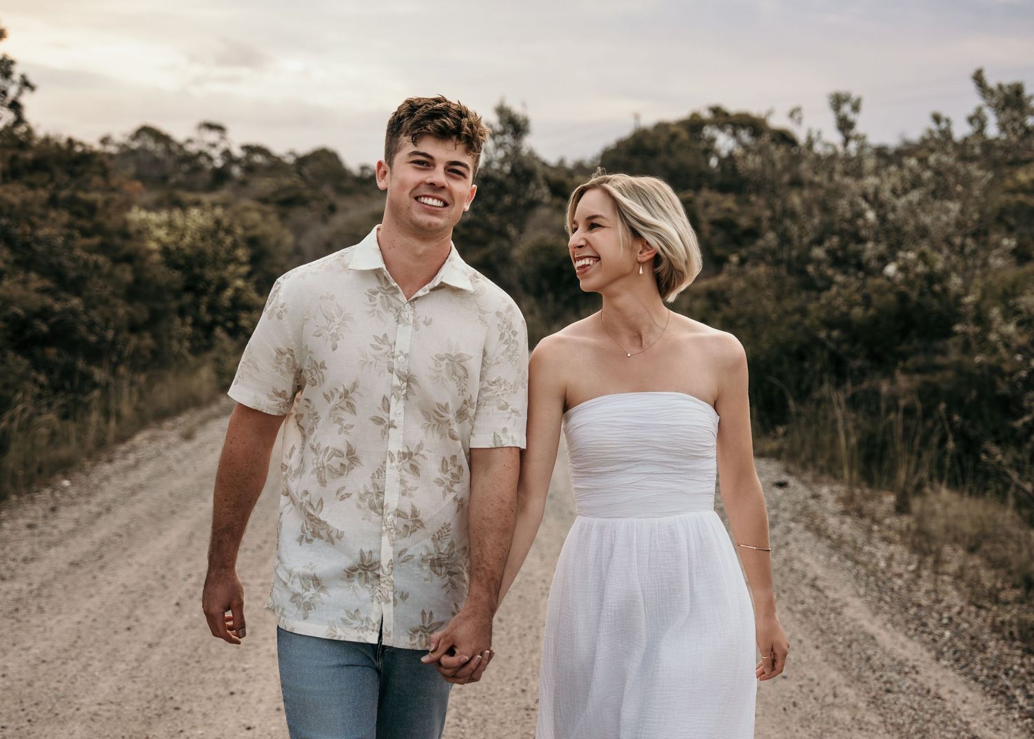 Happy couple holding hands and walking on a dirt road surrounded by trees during golden hour.