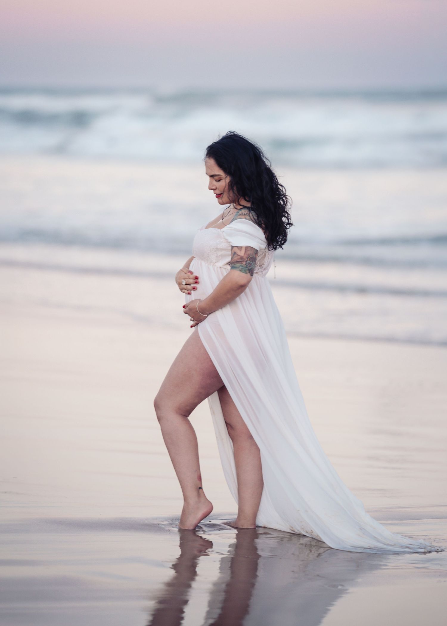 Pregnant woman in white flowing dress holding baby bump on beach at sunset.