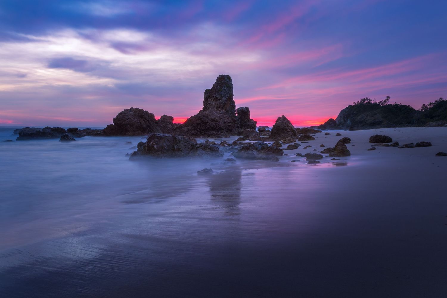 Dramatic purple and pink sunset over rocky sea stacks on a tranquil beach with smooth water reflections.