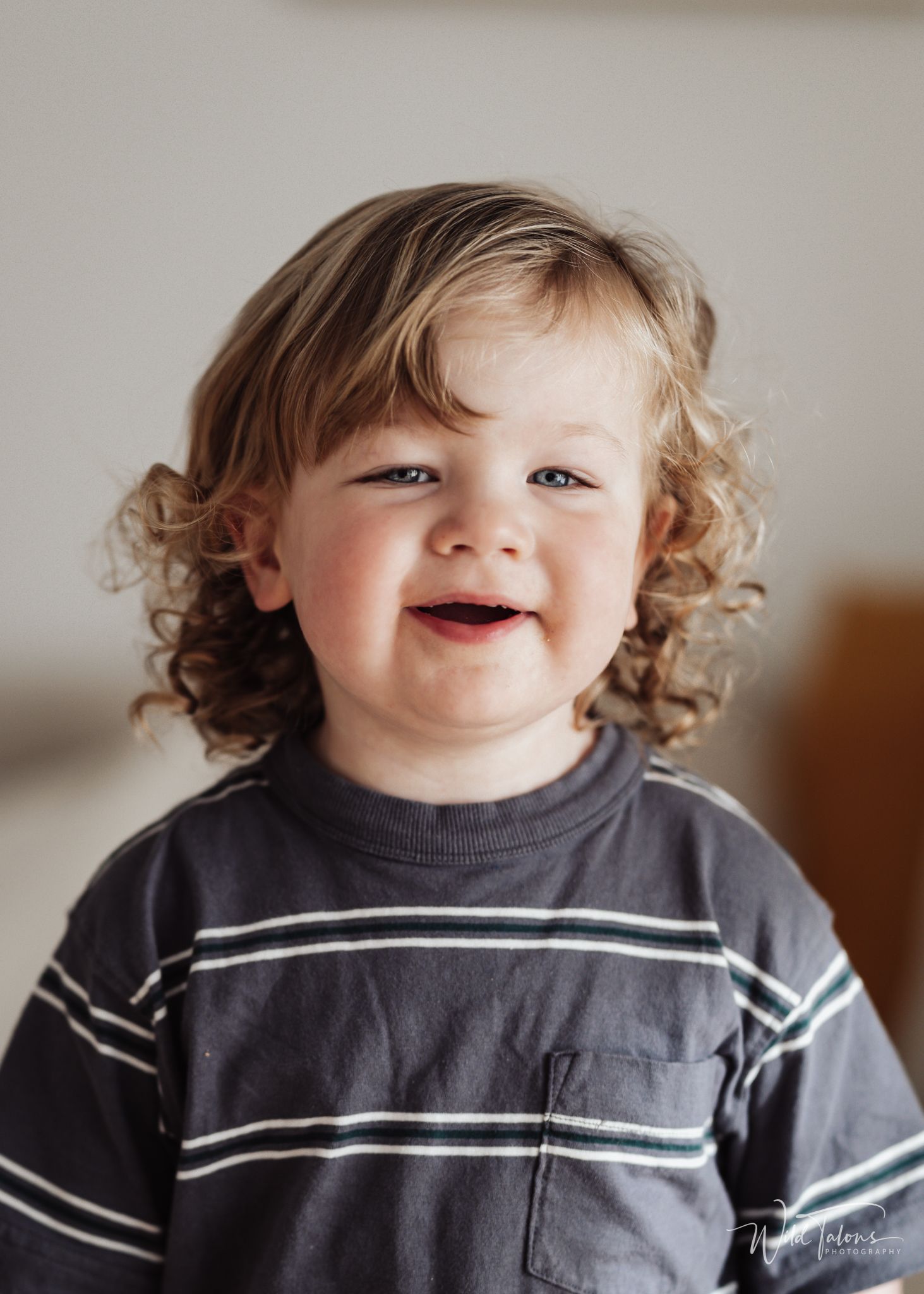Smiling toddler with curly blonde hair wearing a dark striped shirt in a bright indoor setting.