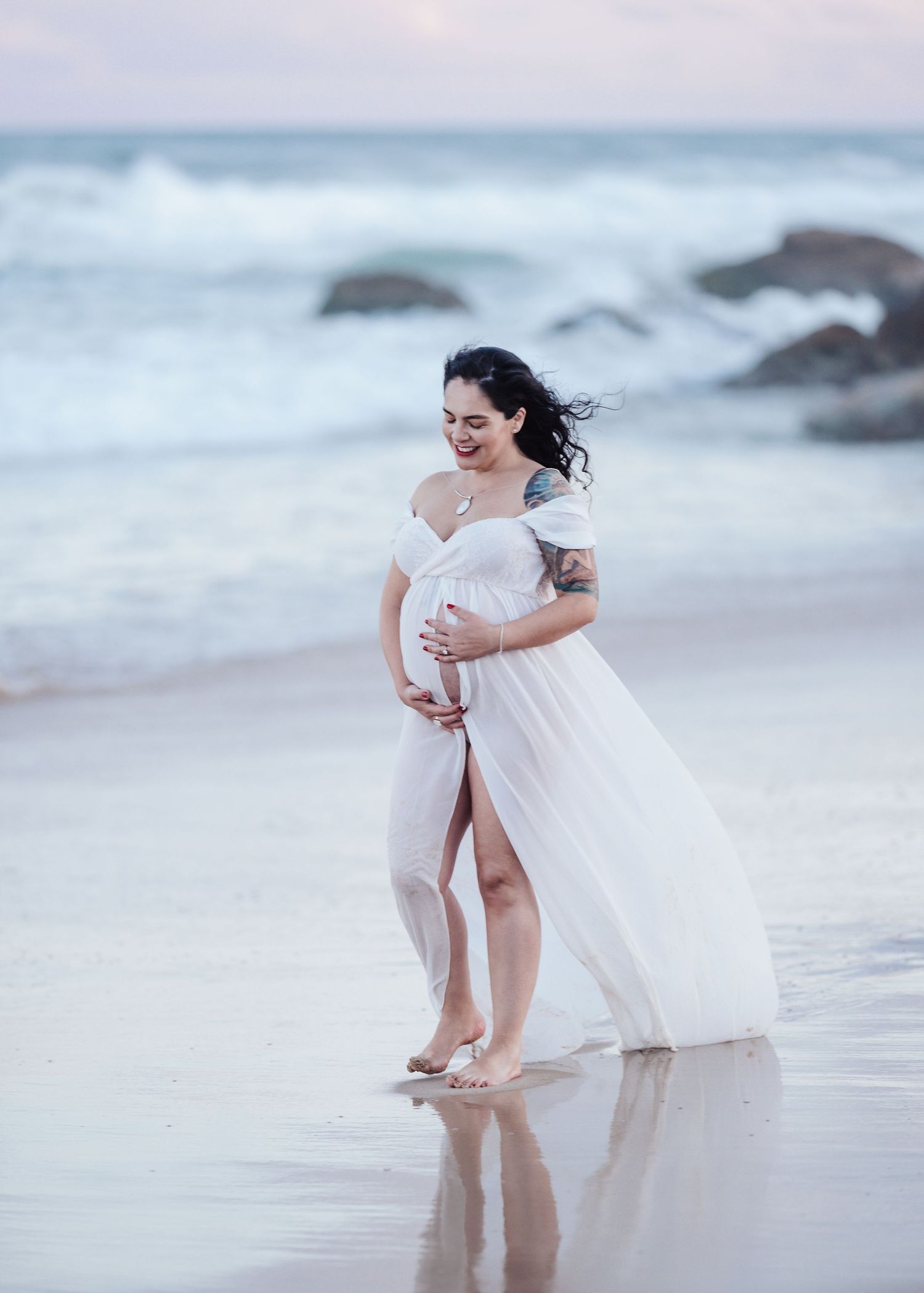 Pregnant woman in white flowing dress standing at ocean's edge laughing during beach maternity shoot.