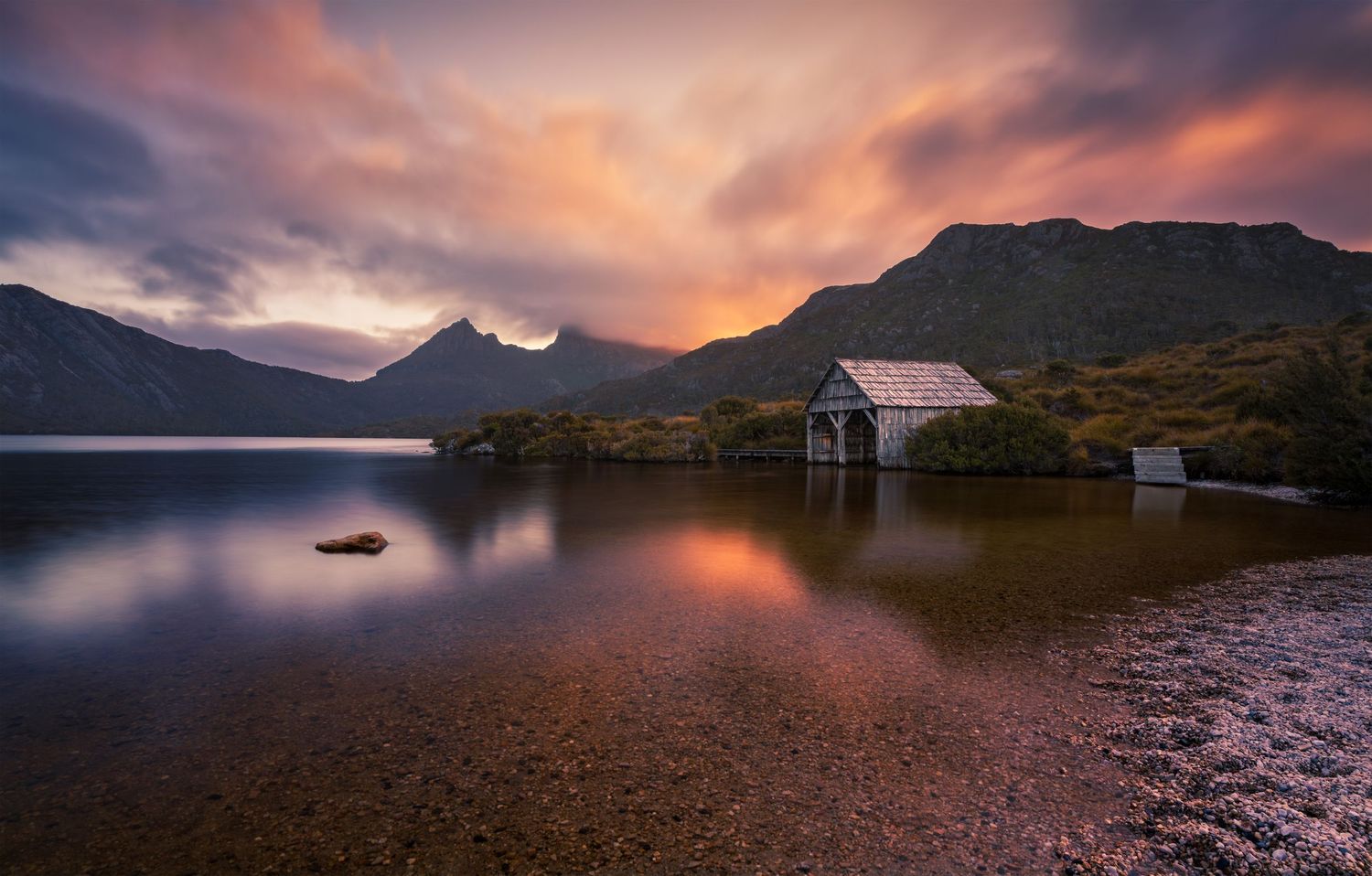 Iconic Dove Lake boathouse glows under a vivid orange and pink sunset sky with Cradle Mountain behind.
