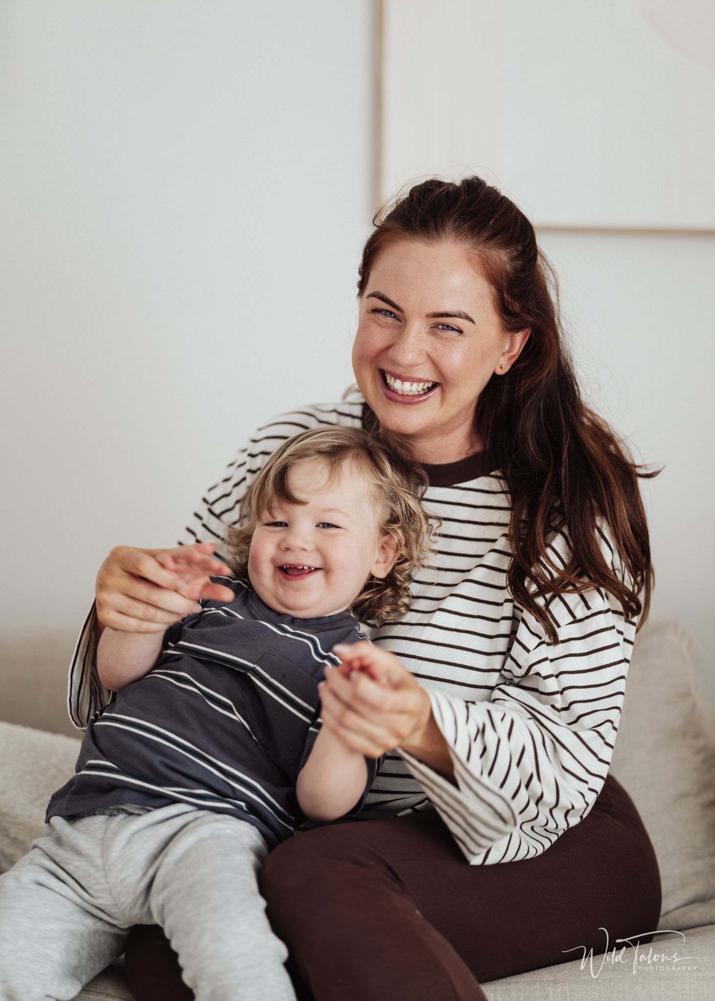 A laughing mother in a striped shirt holds her giggling toddler on a couch in a bright, cozy home setting.