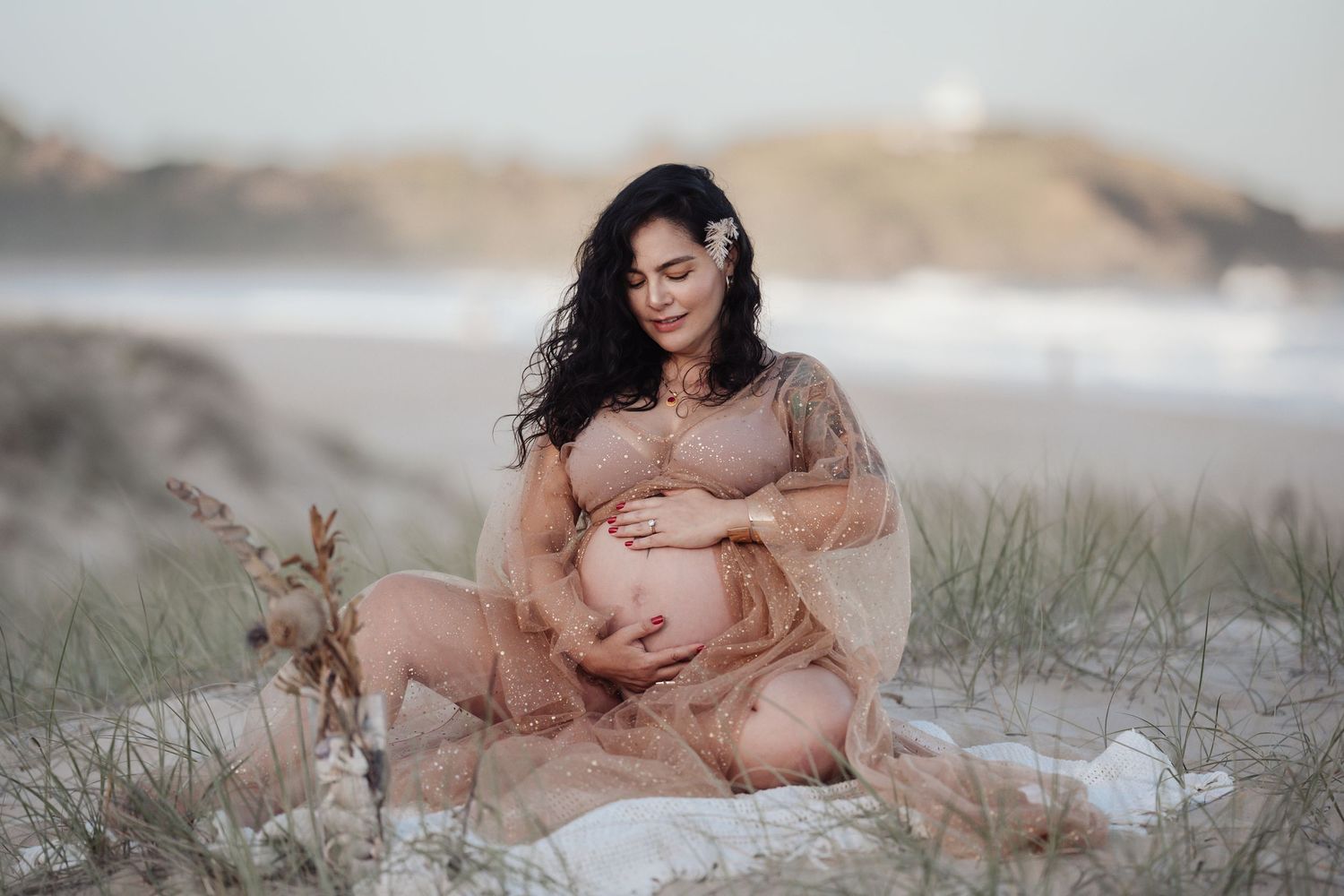 Pregnant woman in sheer blush gown sitting on sand dunes cradling baby bump.