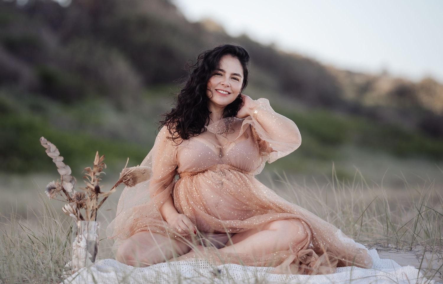 Happy pregnant woman in sheer peach robe sitting on blanket in grassy field.